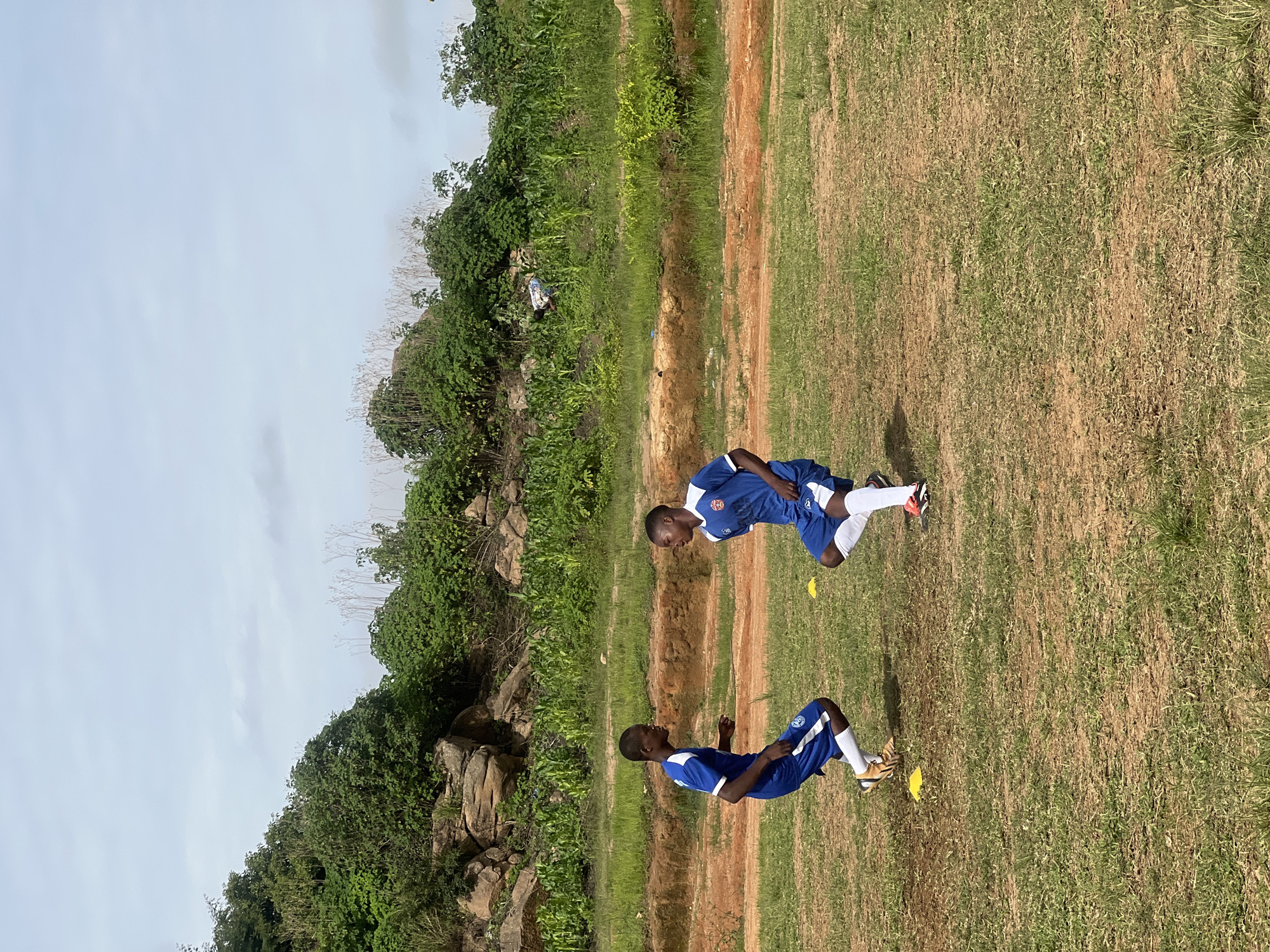 Two young Black males, appearing to be adolescents, are engaged in soccer drills on a grassy field in what is identified as Jos, Nigeria. Both are wearing matching blue and white soccer uniforms, including shorts and jerseys with a crest on the left chest. The male on the left is wearing white socks and gold soccer cleats, while the male on the right is wearing white socks and black and red soccer cleats. They are positioned near yellow training cones on the field. In the background, a steep, rocky incline covered with dense green vegetation and some cultivated crops rises behind a reddish-brown dirt embankment. The sky above is overcast with a light, cloudy haze.