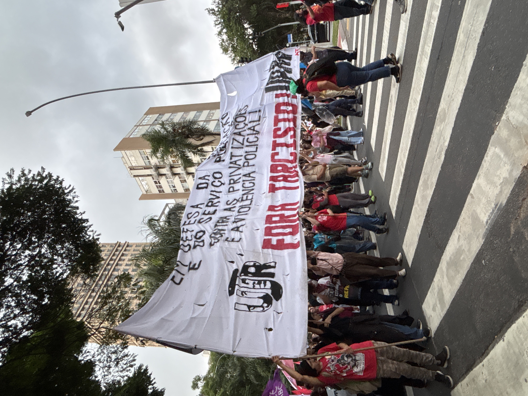 A wide shot depicts a political demonstration taking place in a crosswalk in what is presumably São Paulo, Brazil.
Approximately 25-35 participants are engaged in a line that spans the crosswalk, holding a large, white banner. The banner, written in Portuguese, criticizes privatization and police violence, and bears a slogan that translates to "Out Tarcisio." Tarcisio is presumably a political figure who is unpopular with these demonstrators.
The scene takes place on a paved street with a zebra crosswalk. Buildings and trees can be seen in the background, indicating an urban location. Overcast skies suggest that it may be early morning or late afternoon. A few participants appear to be moving across the crosswalk, while the majority are stationary, seemingly blocking traffic. The crowd is ethnically diverse.