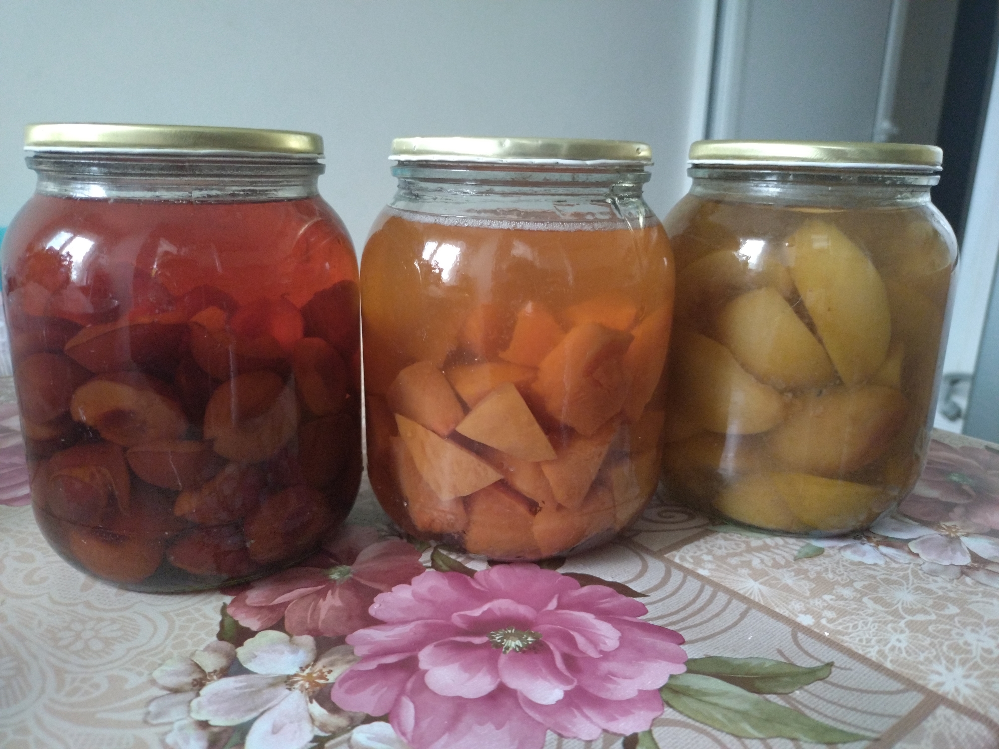 The image shows three glass jars with golden lids, filled with fruit preserves. From left to right, the first jar contains dark red plums in a reddish liquid, the second holds orange fruit pieces (possibly peaches or apricots) in a pale orange liquid, and the third features yellow fruit halves (likely pears or peaches) in a yellow-tinted liquid. The jars are arranged on a tablecloth with a floral pattern, showcasing pink and white flowers with green leaves. The background is a light-colored wall, partially visible. The image appears to be taken indoors. No specific location in Stara Zagora, Bulgaria, can be determined from the image.