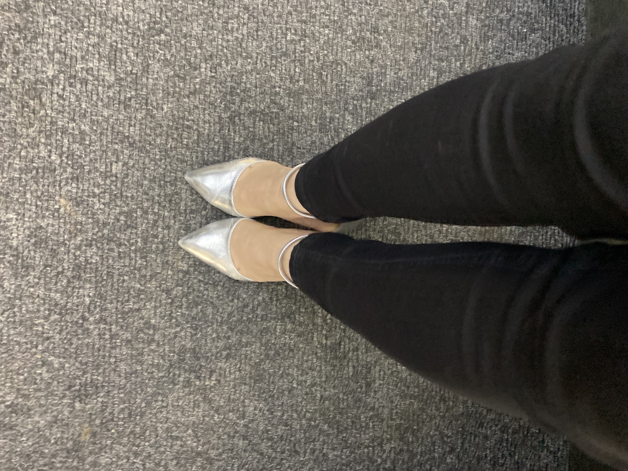 The image shows a person's legs from a top-down perspective, wearing black skinny jeans and silver pointed-toe flats. The person is standing on a gray carpet with a textured pattern. No information suggests that the scene is located in Yekaterinburg, Russia.