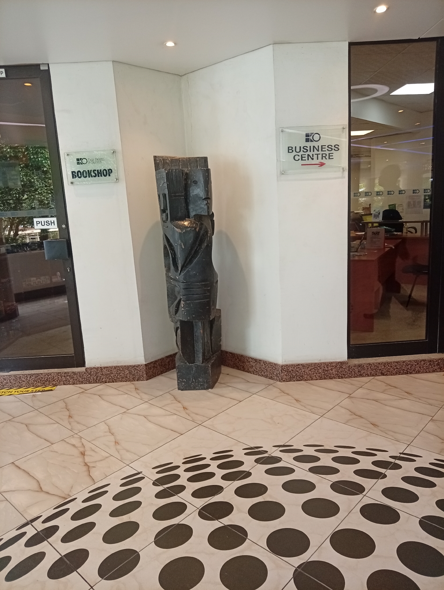 The image shows an interior space, likely a hotel lobby, featuring a decorative sculpture positioned between two glass-door entrances. The doors are labeled "Bookshop" and "Business Centre," indicating nearby facilities. The sculpture is tall, dark, and appears to be made of carved wood or stone. The floor is tiled with a pattern of large black circles on a light background. A yellow "Watch Your Step" sign is visible near the "Bookshop" door. The scene suggests a commercial or hospitality setting. While the image appears to be located in the Eko Hotels and Suites, Itirin, Nigeria, it is not possible to verify this from the image alone.