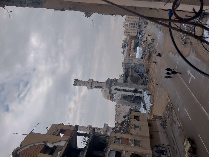 The image depicts a street scene in Khan Yunis, Palestinian Territory, characterized by extensive urban destruction under an overcast sky. In the midground, a large mosque, identified by its prominent minaret and dome, stands significantly damaged; its main structure exhibits substantial collapse, exposed interiors, and widespread debris. The minaret itself appears largely intact at its upper sections but shows signs of weathering or minor damage. Surrounding the mosque are makeshift shelters constructed from corrugated metal.
On both sides of the central street, multi-story buildings are severely damaged, with large sections of walls missing, exposing internal structures, and revealing piles of rubble. Exposed rebar is visible in some areas of the damaged buildings.
The street itself is paved, appears wet in various sections, and features visible white road markings, including a turning arrow and lane lines. Significant accumulations of sand, rubble, and debris are present along the road edges and around the damaged structures.
Several individuals are walking on the street. A group of approximately five people, dressed in dark attire, walks away from the camera in the foreground-midground. Other smaller groups and solitary figures are visible further down the street. On the left side of the street, a two-wheeled utility cart is parked. In the distant background, additional buildings, some appearing less damaged, and a white tent-like structure are visible. The foreground includes a partial view of a dark metal railing, indicating the photograph was taken from an elevated perspective.