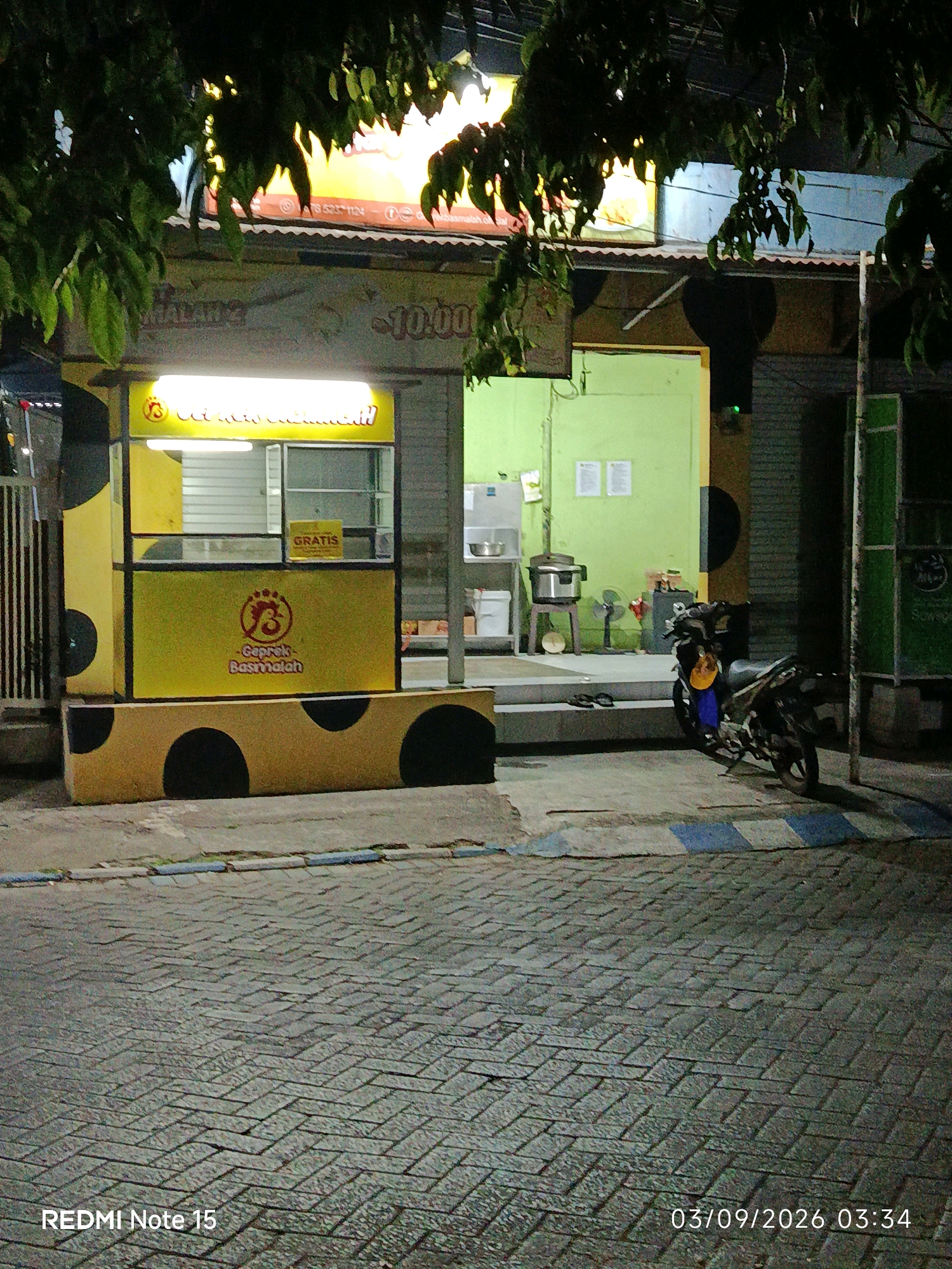 A street-level, medium shot captures a food stall at night in Keboansikep, Indonesia. The stall, brightly lit with a yellow awning and accents, features a sign with the text "Geprek Basmalah" and a chicken logo. A smaller sign displays "GRATIS". The stall has a counter with shutters, and behind it, cooking equipment including a pot is visible. To the right of the stall, a parked motorcycle with a yellow helmet on the seat is illuminated. In the background, a green wall and a metallic structure are present. The foreground shows a cobblestone street with blue and white striped markings. The image is dated "03/09/2026 03:34" and branded "REDMI Note 15".