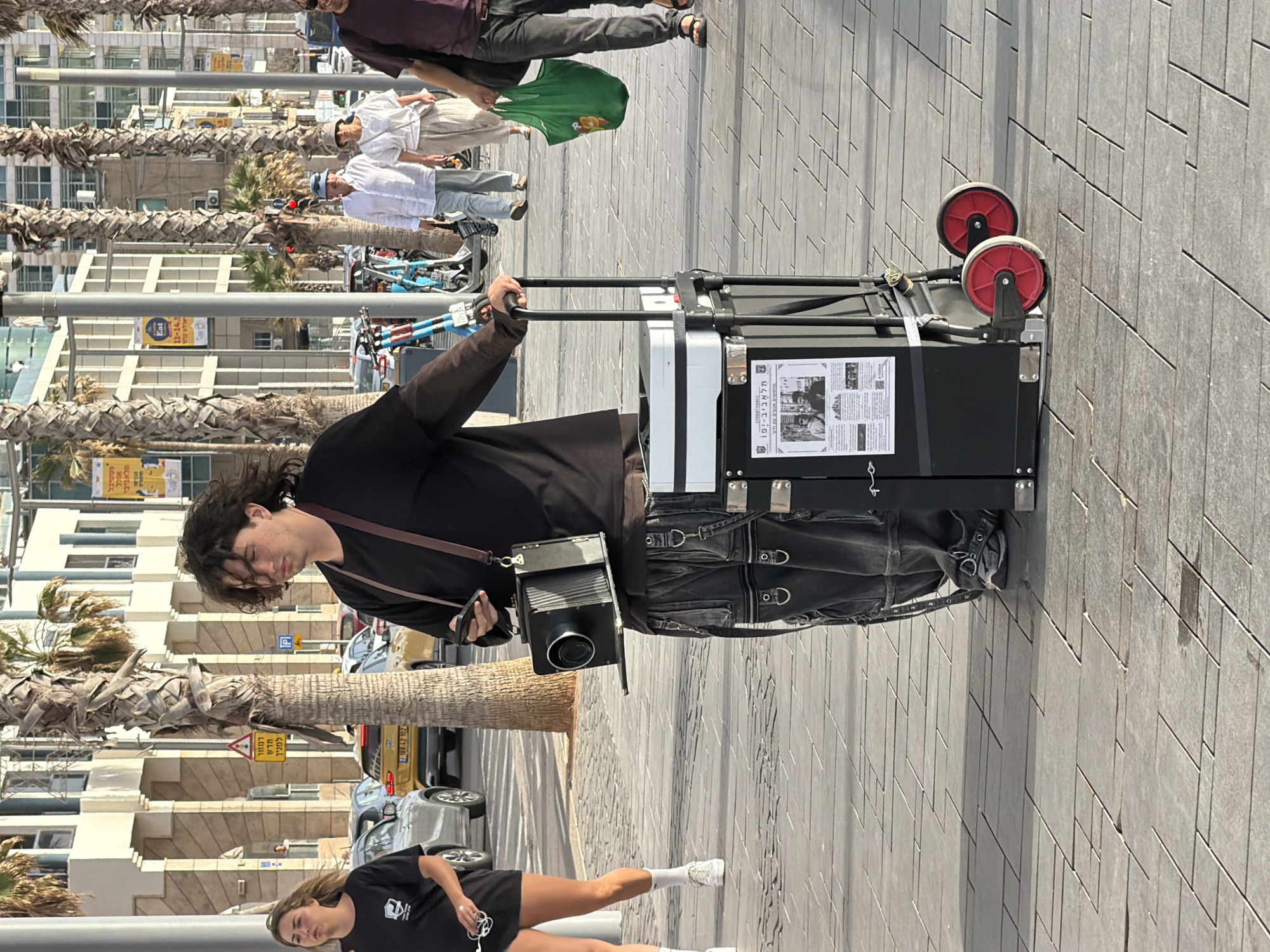 A photographer wearing a black long-sleeved shirt and a camera over his shoulder is shown on a paved street, pulling a black suitcase with red wheels behind him. The suitcase has printed material, possibly a flyer, attached to it. He is walking past palm trees along the sidewalk. In the background, buildings and street signs are visible, including a sign for a festival "Eat 11-14.5." Another sign says "Tel Aviv-Yafo (Jaffa)" along with Hebrew text, indicating the city's location. Additionally, traffic, cars, and other pedestrians are visible. A woman in a black shirt and white socks is walking away from the photographer.