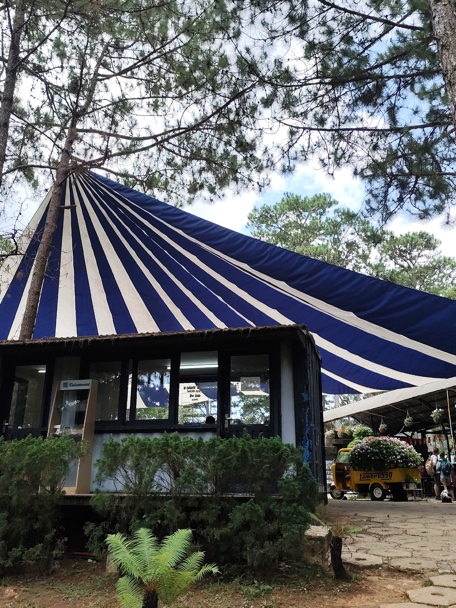 The image shows a building with a distinctive blue and white striped awning. A tree trunk appears to support the awning at one end. In front of the building, a small yellow vehicle with flowers is parked. There are people in the background. The location is Da Lat, Vietnam.