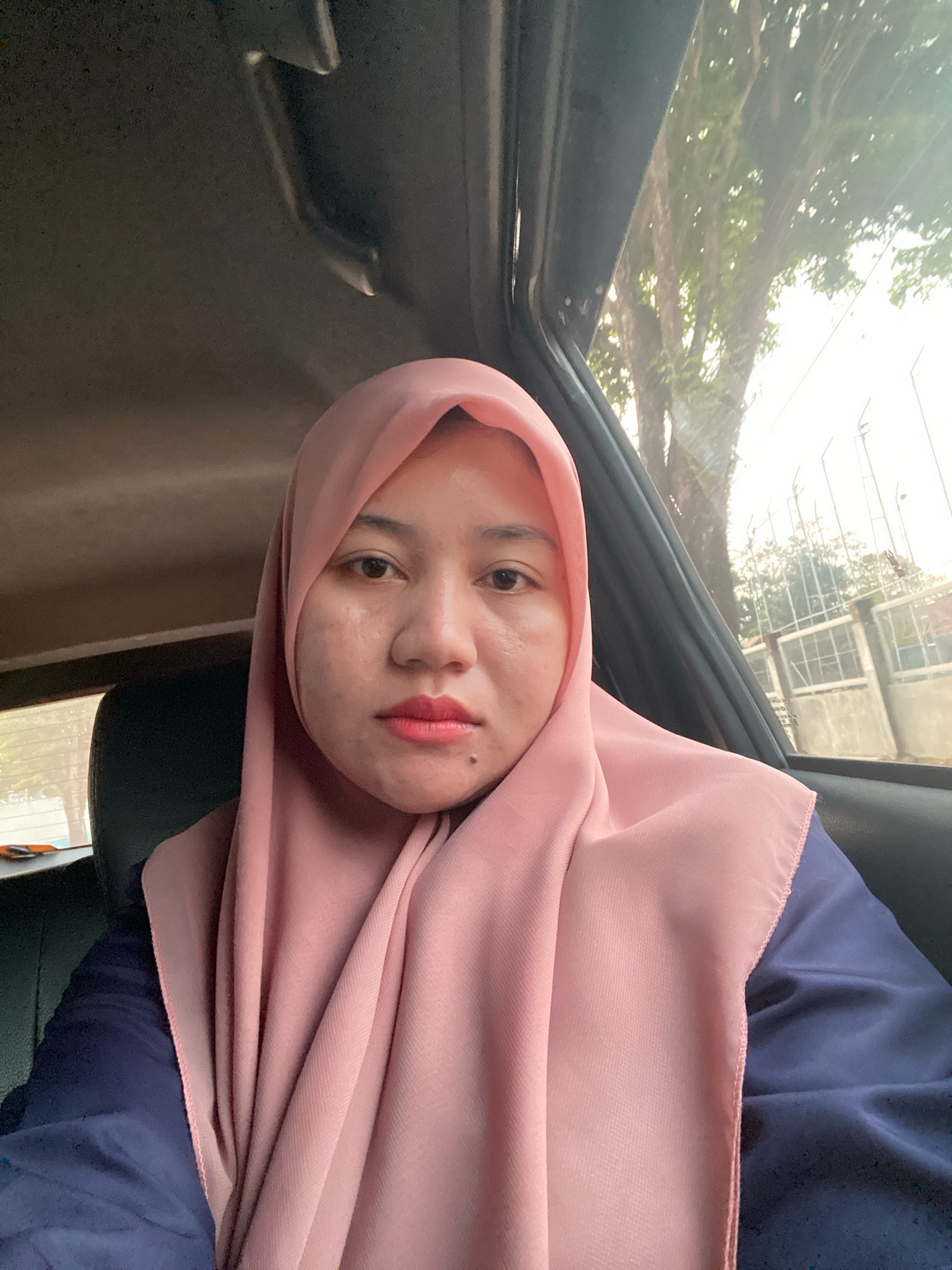 The image is a close-up, selfie-style photograph taken from inside a vehicle. The subject, a woman wearing a pink hijab and a dark blue garment, is looking directly at the camera. Her face is partially visible, with a mole on her chin. The background shows the interior of a car, with a textured headliner, and the exterior through the car windows, depicting trees and a fence. The lighting suggests it is daytime. The scene is consistent with a person taking a photo while traveling in a car within the city of Banjarbaru, Indonesia.