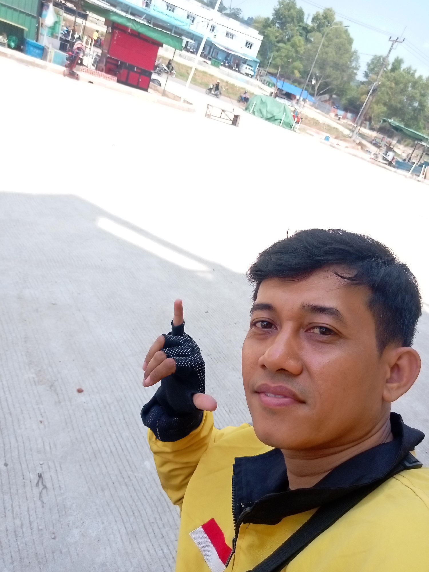 A man with dark hair and brown eyes, wearing a yellow jacket with black trim, a black and white patterned glove on his right hand, and a black strap across his chest, is taking a selfie outdoors. He is pointing his gloved index finger upwards. The background shows a paved area with concrete lines, a building with a red stall and a green awning, and other buildings and trees. A white line is visible on the pavement in the upper right portion of the image. The scene appears to be a public area in Batam, Indonesia, possibly a market or a service area.