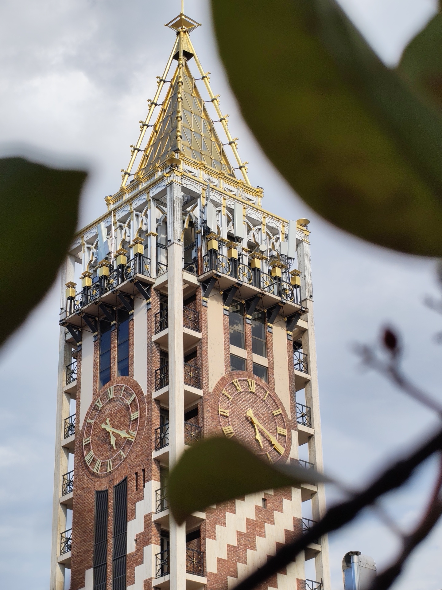 A tall clock tower, characteristic of Batumi, Georgia, stands against a cloudy sky. The tower is constructed with red brick and cream-colored accents, featuring large circular clock faces with Roman numerals and gold hands. Decorative balconies with black railings and gold accents adorn the upper levels, which are topped by a golden, pyramid-shaped spire. Foliage obscures parts of the foreground, hinting at an outdoor setting.