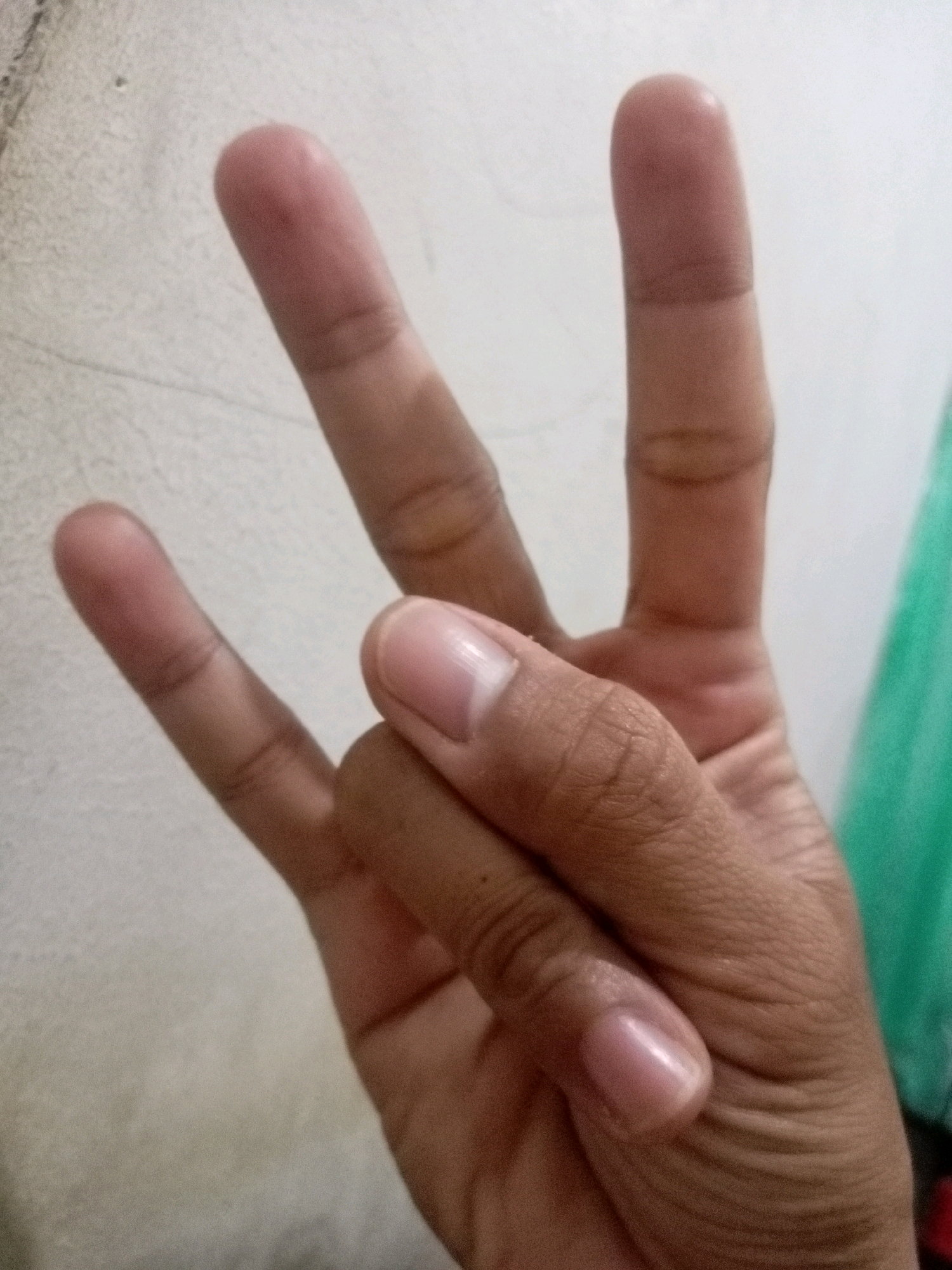 The image shows a close-up of a person's hand. Three fingers are visible, extended upwards, with the thumb tucked under. The index finger and middle finger are raised, forming a "V" shape. The ring finger is also extended. The background is a light-colored, textured wall. The hand is positioned in the foreground, with the fingers taking up most of the frame. The lighting appears to be indoor. There is no identifiable information in the image that would suggest a location in Batam, Indonesia, or any specific urban context. The image is purely a depiction of a hand gesture.