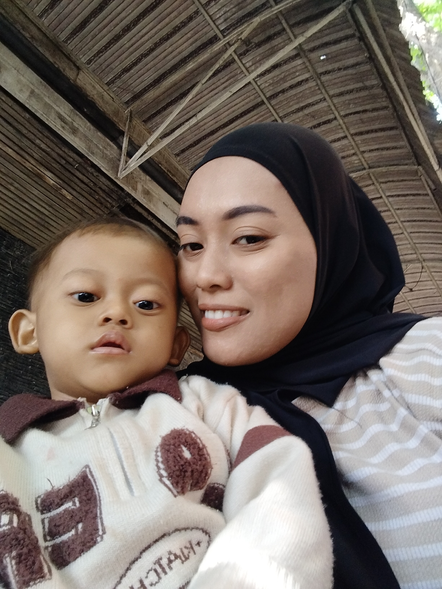 A close-up, low-angle selfie shows a woman wearing a black hijab and a beige and white striped shirt, holding a young child. The woman, on the right, has her face turned towards the camera, smiling with her teeth showing. Her left eye and forehead are visible. The child, on the left, has a neutral expression and is looking directly at the camera. The child is wearing a light-colored sweater with brown accents, including a zipper on the front. The background consists of a rustic, wooden roof structure, suggesting an outdoor or semi-outdoor setting, possibly in a rural area or a less developed part of Kecamatan Pati, Indonesia. The lighting is natural and appears to be daytime.