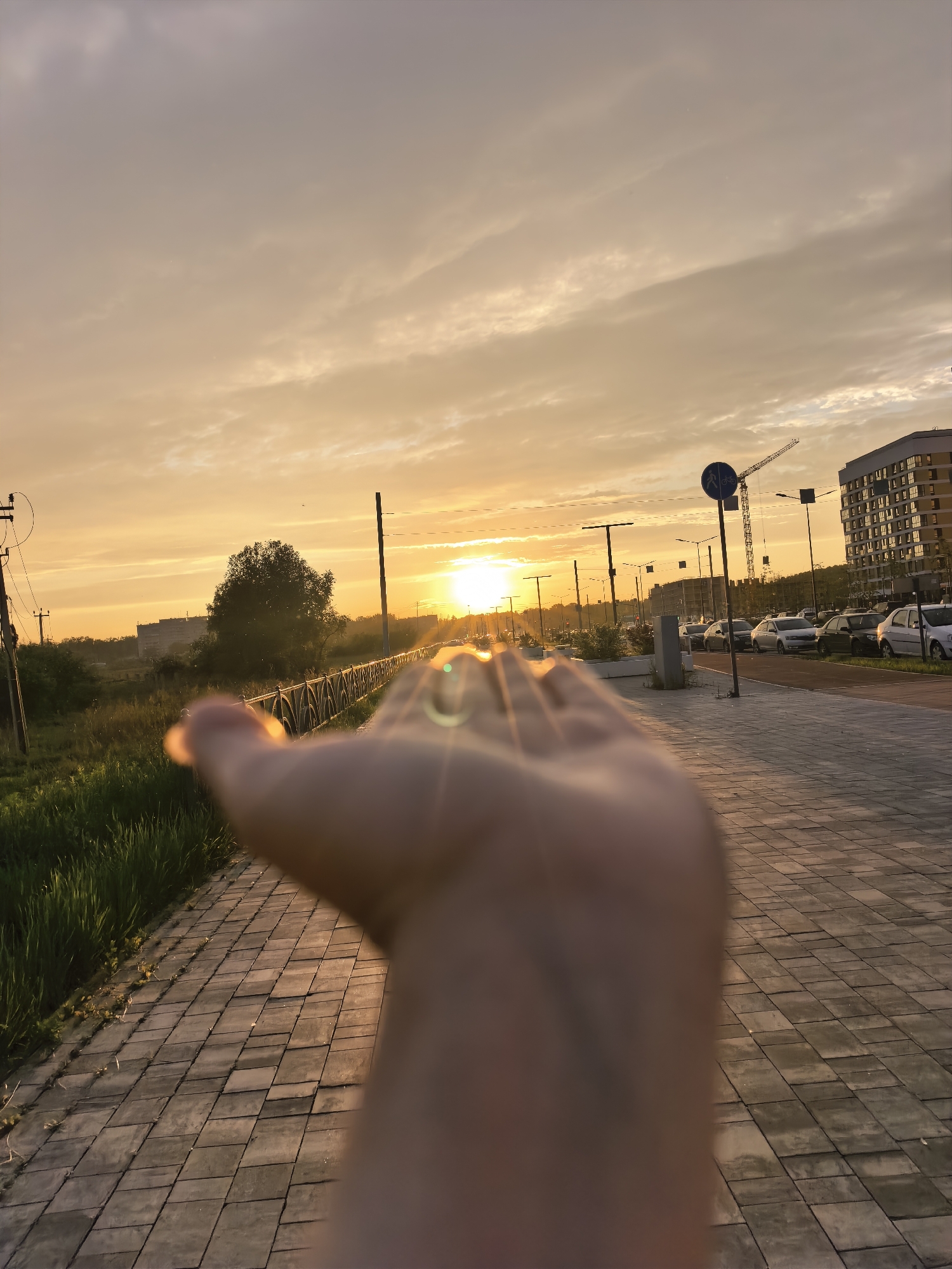 A first-person perspective captures a sunset in an urban environment. The foreground is dominated by a blurred human hand, extended towards the camera. The sun, a bright orb, is visible in the mid-ground, casting a warm glow and creating lens flares. The background shows a paved walkway, a grassy verge with a fence, streetlights, parked cars, a residential building, and a construction crane. The sky is filled with soft clouds tinged with orange and yellow from the setting sun.