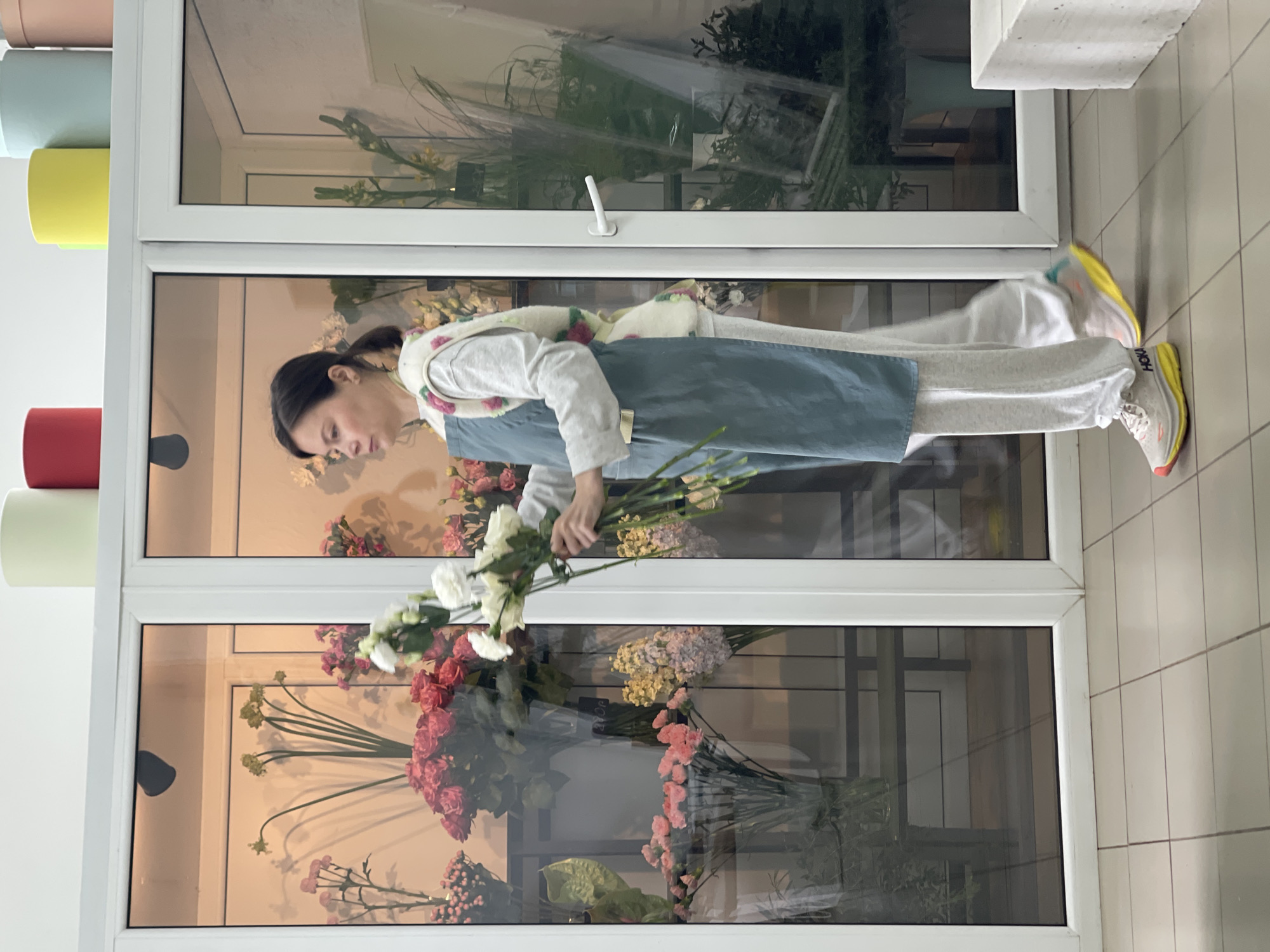 The image depicts a woman engaged in floristry within what appears to be a flower shop or studio. She stands before multiple large window panes with white frames, which display numerous colorful flower arrangements.
The woman has dark hair pulled back and is wearing a light grey long-sleeved top, a white vest with pink, red, and green floral patterns, and a light blue/grey apron. Her lower attire consists of light grey sweatpants and white athletic shoes with bright yellow soles and red accents, featuring the brand name "HOKA" on the side. She is holding a large bunch of white flowers, focused on arranging or preparing them.
Through the window panes behind her, various bouquets are visible, including red, pink, yellow, and light purple flowers, along with green foliage. These reflections create a sense of depth and abundance within the floral display. To the left of the frame, a white shelving unit or wall fixture holds stacked cylindrical containers in different colors: red, light green, yellow, light blue, light pink, and beige/brown. The floor is tiled with light-colored square tiles. The lighting is bright and even, suggesting an indoor setting with ample illumination, possibly from natural light. The scene conveys a professional and artistic environment centered around floral design.