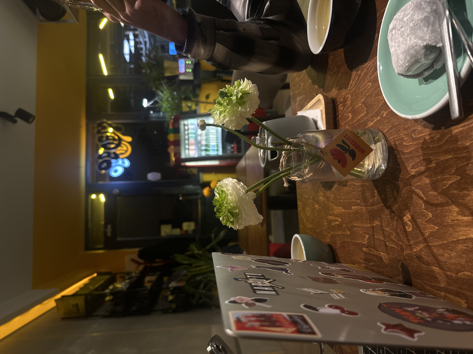The image captures a scene at a cafe in Yerevan, Armenia. The camera is focused on a wooden table, on which are several items including a laptop covered in stickers, a small vase with two white flowers, a cup of tea, and a plate with a gray mochi dessert. A person wearing a plaid shirt is partially visible at the top of the frame. In the background, through a window, there is a view of the street at night, illuminated by the cafe's neon sign and lights from nearby buildings, including a vending machine.