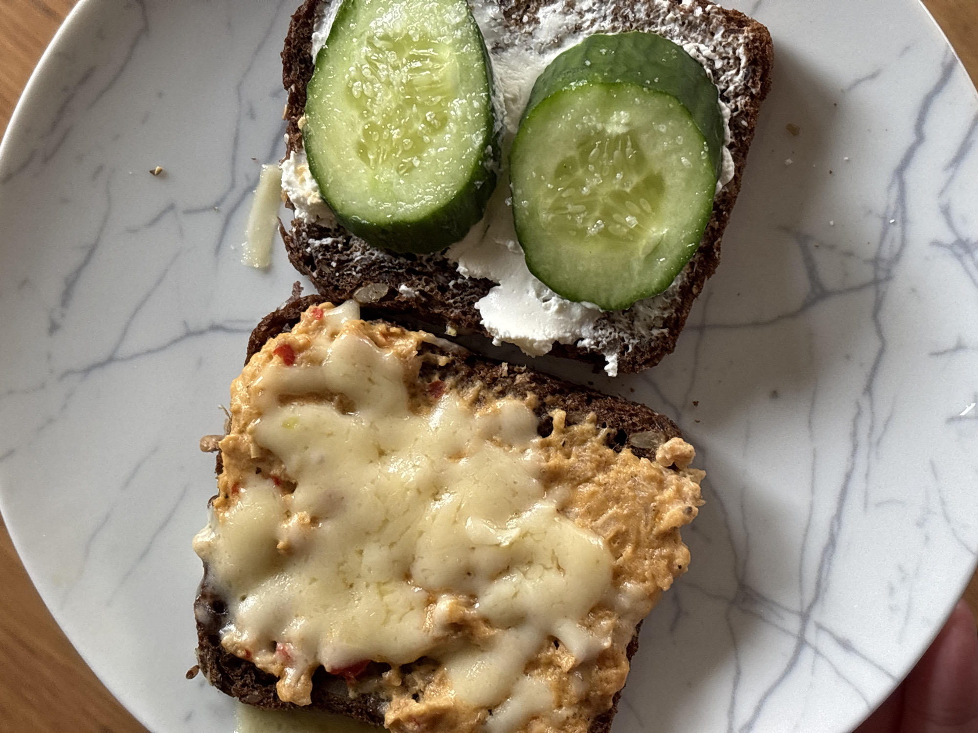 The image shows two open-faced sandwiches on a marble-patterned plate. The top sandwich consists of a slice of dark bread spread with a white creamy substance, topped with two slices of cucumber. The bottom sandwich is made of the same dark bread, topped with a chunky spread and covered with melted cheese. The plate appears to be resting on a wooden surface. The image provides no information related to the location in the city of Мещёры, Russia, or any people or interactions.