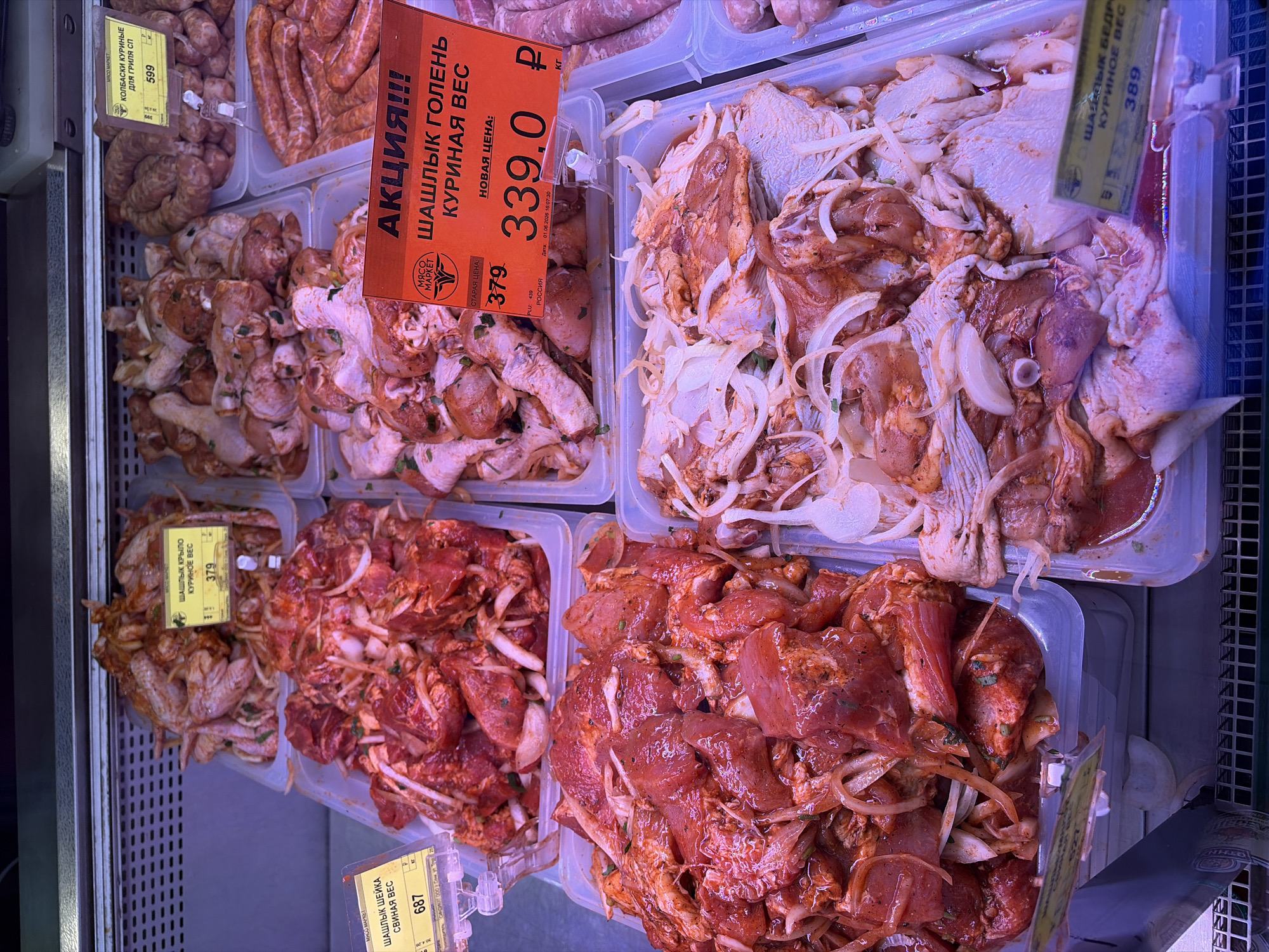 This image captures an overhead view of a refrigerated display case inside a meat market, likely in Sochi, Russia. The scene is filled with various types of raw, marinated meat products packaged in clear plastic containers, ready for sale.
The primary subjects are the diverse cuts of meat. In the top left, coiled chicken sausages are visible, marked with a yellow price tag displaying "КОЛБАСКИ КУРИНЫЕ ДЛЯ ГРИЛЯ СП" (Chicken sausages for grill SP) and a price of "599". Adjacent to these are containers of marinated chicken drumsticks (goleni). A prominent orange price tag advertises a "АКЦИЯ!!!" (SALE!!!) for "ШАШЛЫК ГОЛЕНЬ КУРИНАЯ ВЕС" (Chicken Leg Skewers, by weight), showing an "СТАРАЯ ЦЕНА: 379" (Old Price: 379) and a "НОВАЯ ЦЕНА: 339.0 Р" (New Price: 339.0 R). This label also includes "МЯСО МАРКЕТ" (Meat Market), "РОССИЯ" (Russia), and a date "01.06.2026 16:07:30".
To the right, another container holds marinated chicken pieces, possibly thighs or breasts ("ШАШЛЫК БЕДР КУРИНОЕ ВЕС" - Chicken Thigh Skewers, by weight), with visible slices of onion mixed into the marinade, priced at "389".
The bottom row features more marinated meats. On the left, chicken wings are seen, labeled "ШАШЛЫК КРЫЛО КУРИНОЕ ВЕС" (Chicken Wing Skewers, by weight) for "379". The largest container on the bottom right holds marinated pork neck, identified as "ШАШЛЫК ШЕЙКА СВИНАЯ ВЕС" (Pork Neck Skewers, by weight) and priced at "687". All the marinated meats feature thinly sliced white onions and green herbs, suggesting they are prepared for grilling or barbecuing (shashlyk).
The bright lighting suggests an indoor retail environment, and the clear containers allow customers to easily view the products. The overall activity depicted is the retail display and sale of pre-marinated meats in a supermarket or specialized meat store.