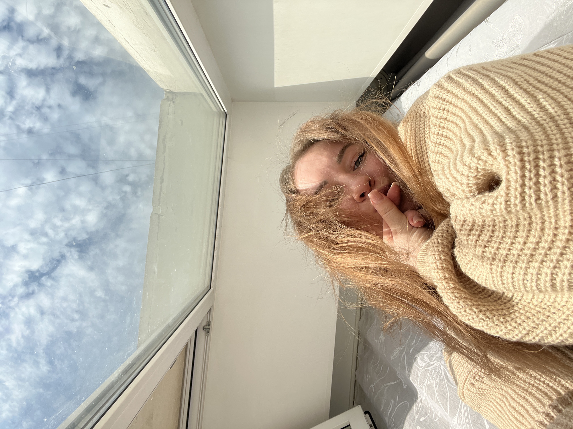 A female individual is depicted in a close-up, high-angle selfie within an indoor setting in Chaikovsky, Russia. The subject, with long, light brown/auburn hair disheveled around her face, is lying on a bed covered with white or light-grey patterned bedding. She wears a light beige, chunky knit sweater. Her right hand, with manicured nails, is positioned with fingers touching her mouth. Her eyes, partially visible, appear light in color as she looks directly at the camera. Above her, a large, rectangular skylight occupies the upper half of the frame, reflecting a clear blue sky with scattered white cumulus or stratocumulus clouds. Faint, thin dark lines, consistent with overhead power lines or cables, are visible against the sky reflection in the upper portion of the skylight. The interior walls surrounding the skylight and behind the subject are uniformly white, illuminated by bright natural light entering through the skylight. The skylight frame appears to be a light-colored metal or PVC.