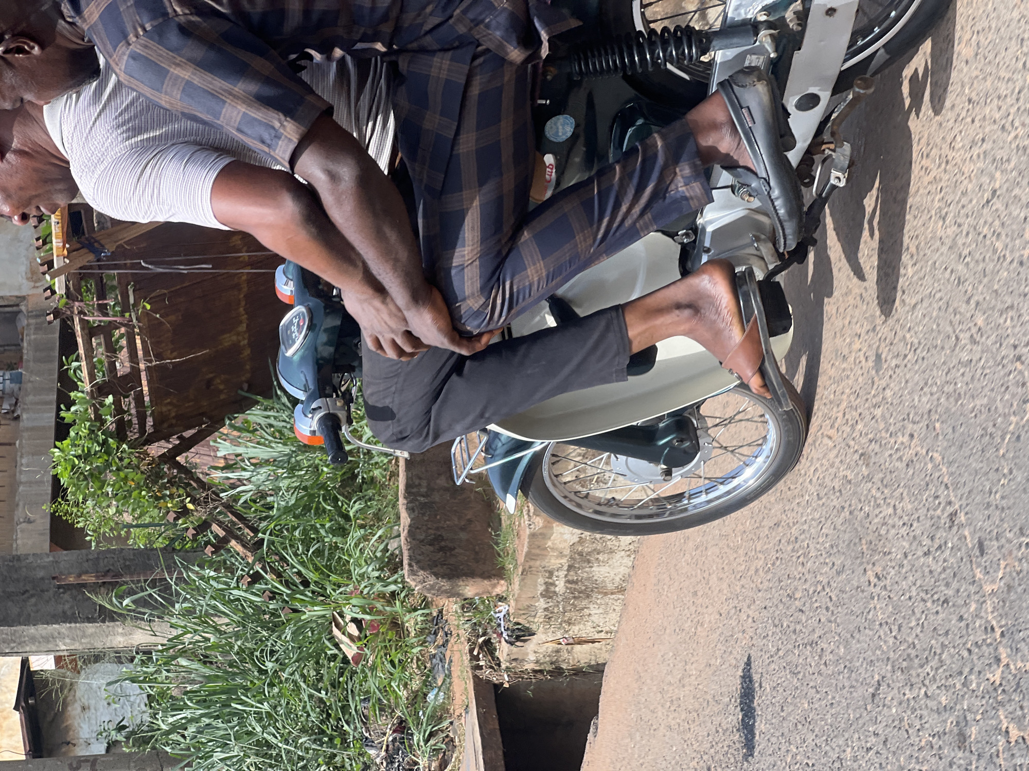 The image depicts two men on a motorcycle, captured from a side angle on a sunny day in Nkpor, Nigeria. The scene takes place outdoors on what appears to be a paved or dirt road beside an unpaved shoulder with abundant vegetation.
The man in the front, presumably the driver, wears a light-colored, possibly white or cream, ribbed short-sleeved t-shirt. His back and right arm are visible. The passenger, seated behind him, wears a dark blue and brown plaid long-sleeved shirt and dark trousers. He is holding onto the driver, indicating they are either riding or poised to ride the motorcycle.
The motorcycle itself is a utility model, possibly a Honda Cub, with a dark body and chrome accents on the wheels and suspension components. A small red sticker on the motorcycle's body, below the seat, features the text "GUD". The passenger's left foot is clad in a black loafer, with faint white text "DEE" visible inside. His right foot wears a brown sandal or flip-flop.
The background shows lush green vegetation, including tall grasses and shrubs, growing along the roadside. There are also concrete structures and a rusty metal fence or gate among the greenery. A visible drainage ditch or gutter runs parallel to the road. The road surface is light-colored, and prominent shadows are cast on it, indicating bright sunlight, likely mid-morning or afternoon, and clear weather. The overall setting suggests a vibrant yet somewhat rugged semi-urban or roadside environment typical of the region.