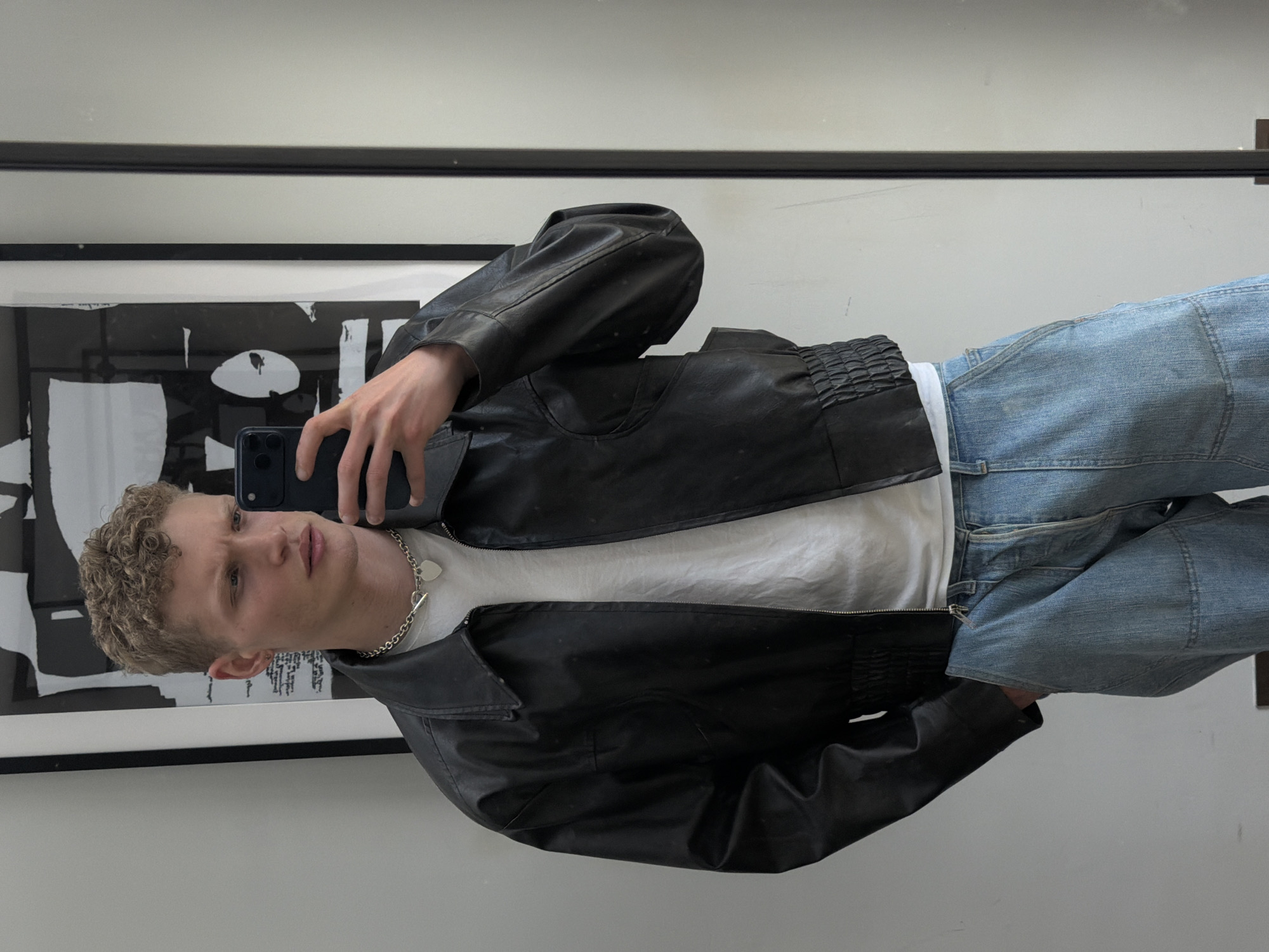 A young man is captured in a mirror selfie, standing upright and looking directly into the camera. He has light-colored, curly hair and fair skin, with a neutral expression. He is wearing a black leather bomber jacket, which features a gathered or ruched elasticated hem and cuffs, over a plain white crew-neck t-shirt. A silver chain necklace with a heart-shaped pendant is visible around his neck. He is also wearing light-wash denim jeans, which appear to have a relaxed fit. His right hand holds a dark-colored smartphone, used to take the picture.
The setting is indoors, likely a room with a large mirror against a plain wall. Reflected in the mirror behind the man is a framed piece of abstract art, characterized by bold black and white shapes. No specific time of day or outdoor weather conditions can be determined from the image. There is no visible text in the photograph.