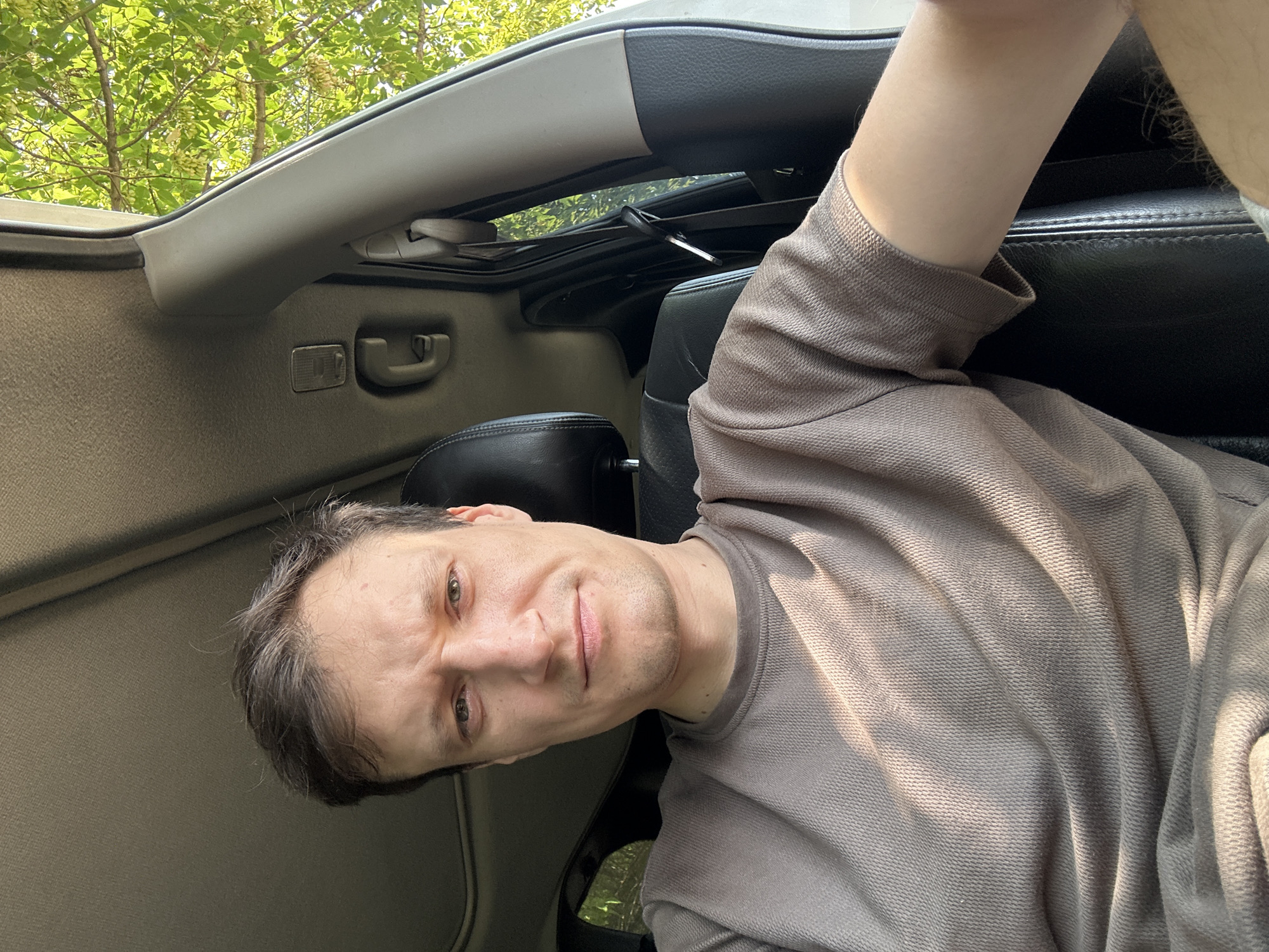 A man is sitting in the driver's seat of a car, looking at the camera. He has short brown hair and is wearing a taupe t-shirt. The interior of the car is visible, including the headrest of the passenger seat and the car's sunroof. Outside the car, green trees are visible, indicating an outdoor setting. The image appears to have been taken during the daytime, with sunlight streaming into the car. The location in Makiv, Ukraine is not specified in the image or its context.