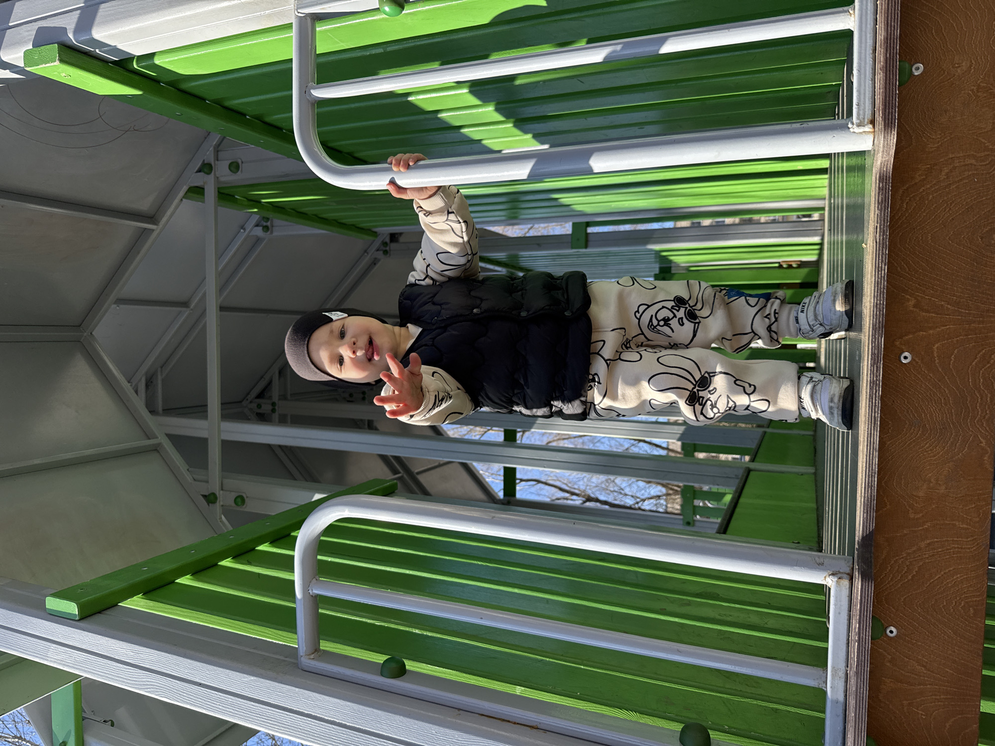 A young child, wearing a dark beanie, black vest, and patterned beige sweat suit, is hanging from a white and green metal jungle gym in Rusanovo, Russia. The child is smiling and holding up a "peace" sign with their left hand. The jungle gym is composed of green corrugated metal and white metal bars. A gray structure is partially visible in the background. Trees are visible through the bars of the jungle gym.