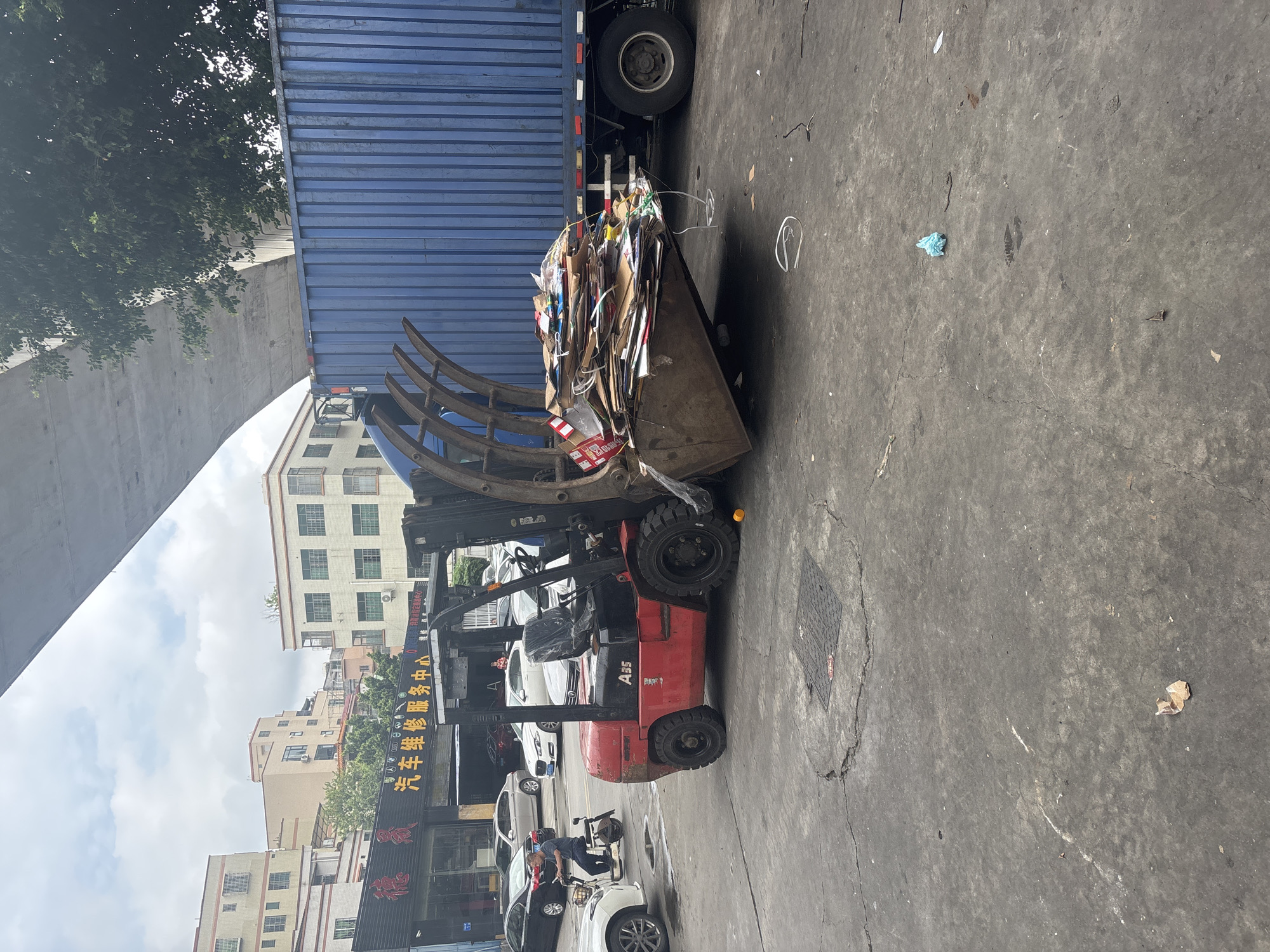 In an outdoor industrial area, likely in the Nanhai District of China, a red forklift with a grapple attachment is positioned next to a blue shipping container. The forklift is in the process of loading or unloading a large pile of cardboard and other debris into the container, with some of the material spilling onto the concrete ground. A person is riding a scooter on the left side of the image, and several cars are parked in the background in front of a row of buildings. The scene is under a large overpass, with trees visible behind the container. The ground is cracked and worn concrete, with some scattered debris.