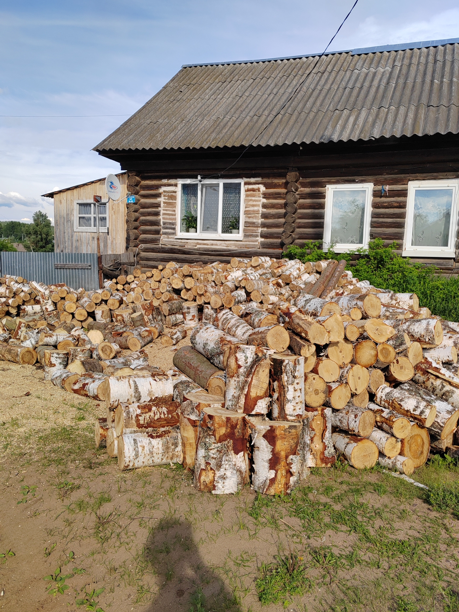 This media file captures a rural or semi-rural scene featuring a traditional wooden log house with an attached wooden plank structure, set against a partially cloudy blue sky. In the foreground, a large pile of freshly cut firewood, predominantly birch logs with their characteristic white and dark bark, is neatly and semi-neatly stacked on the ground. The ground is a mix of bare earth, patches of green grass, and wood chips or sawdust.
The main house has a dark, corrugated roof, possibly made of asbestos cement sheets, and several white-framed windows, some adorned with lace curtains. An attached lighter-colored wooden structure is visible to the left, also with a white-framed window. A small, white satellite dish is mounted on the side of this structure. A corrugated metal fence runs behind the firewood pile on the left, and distant trees are visible beyond it. Green bushes grow at the base of the main house on the right.
The visible text includes "ТЕЛЕКАРТА ТВ" (Telekarta TV) on the satellite dish, and a small blue sign nearby with "ТРЙКОЛОР ТВ" (Trikolor TV) and numbers "812 88 88". Another small blue rectangular sign on the main house displays "21" and possibly "2А" or "2Д", likely a house number.
The scene depicts preparation for heating, suggesting a location where wood is a primary fuel source, possibly in a colder climate. The time of day is bright, likely morning or afternoon, indicated by clear daylight and visible shadows on the ground from a person standing to the right. The overall impression is one of practical rural living.