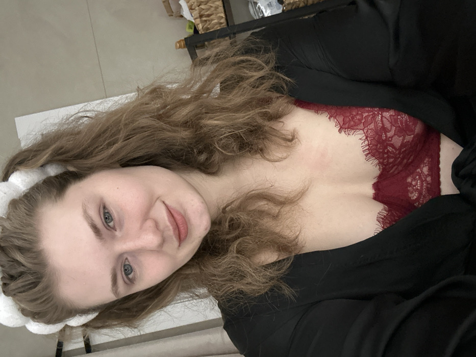 A woman lies on her side, gazing at the camera. She has wavy brown hair, fair skin, and blue eyes. A white spa headband sits atop her head. She is wearing a black robe open to reveal a burgundy lace bra. The background is blurred, showing a light-colored floor and a shelf with baskets on top. The image is slightly tilted.