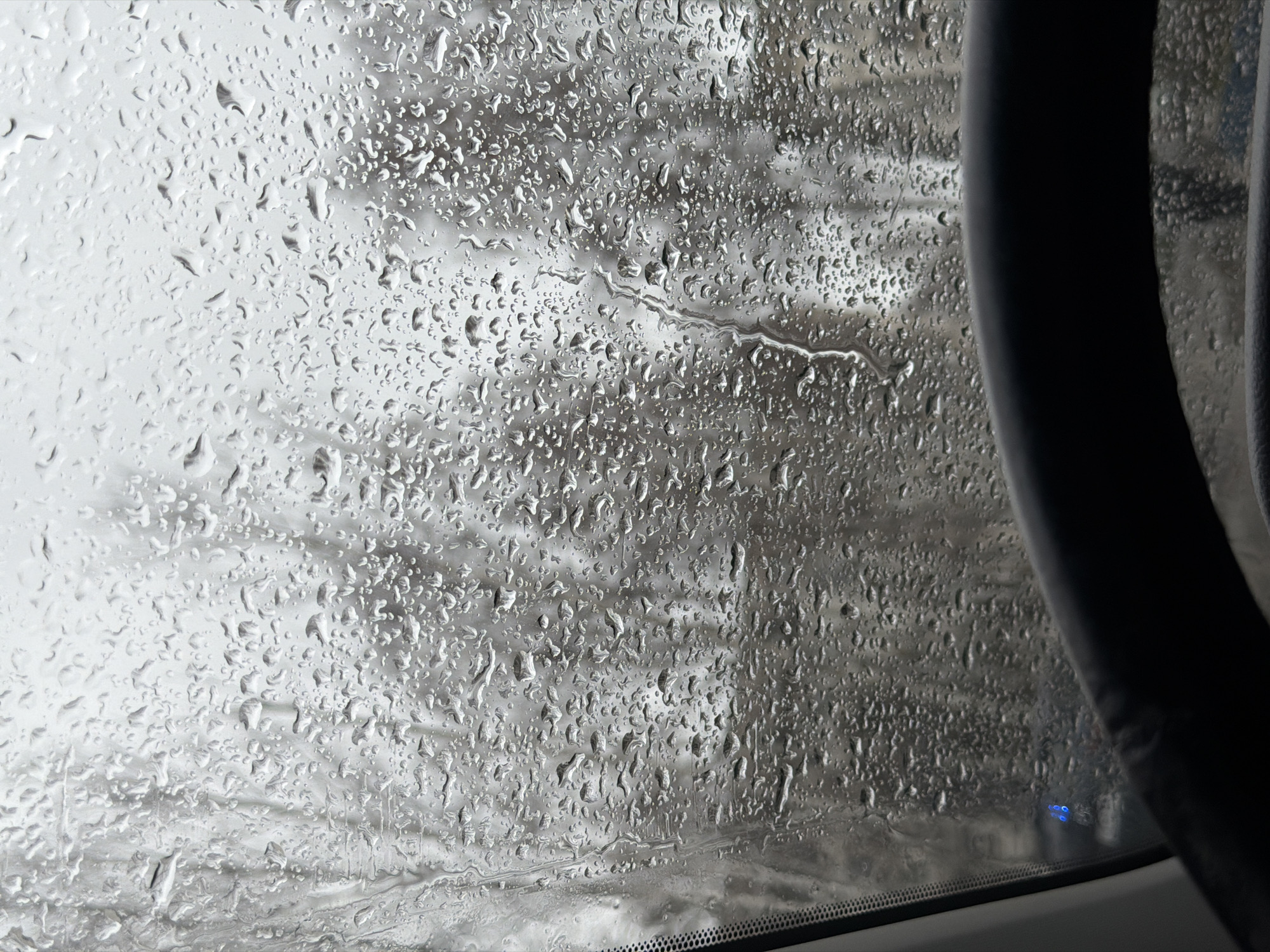 The image shows a view through a car window covered in raindrops. On the right side of the image, a portion of the car's interior, possibly the steering wheel, is visible. The windshield has accumulated numerous water droplets, obscuring the view outside. The background appears to be an indistinct gray landscape, possibly trees or buildings. It is impossible to confirm the location as Chaikovsky, Russia from the visual content alone. There are no discernible people, actions, or interactions depicted in the image.