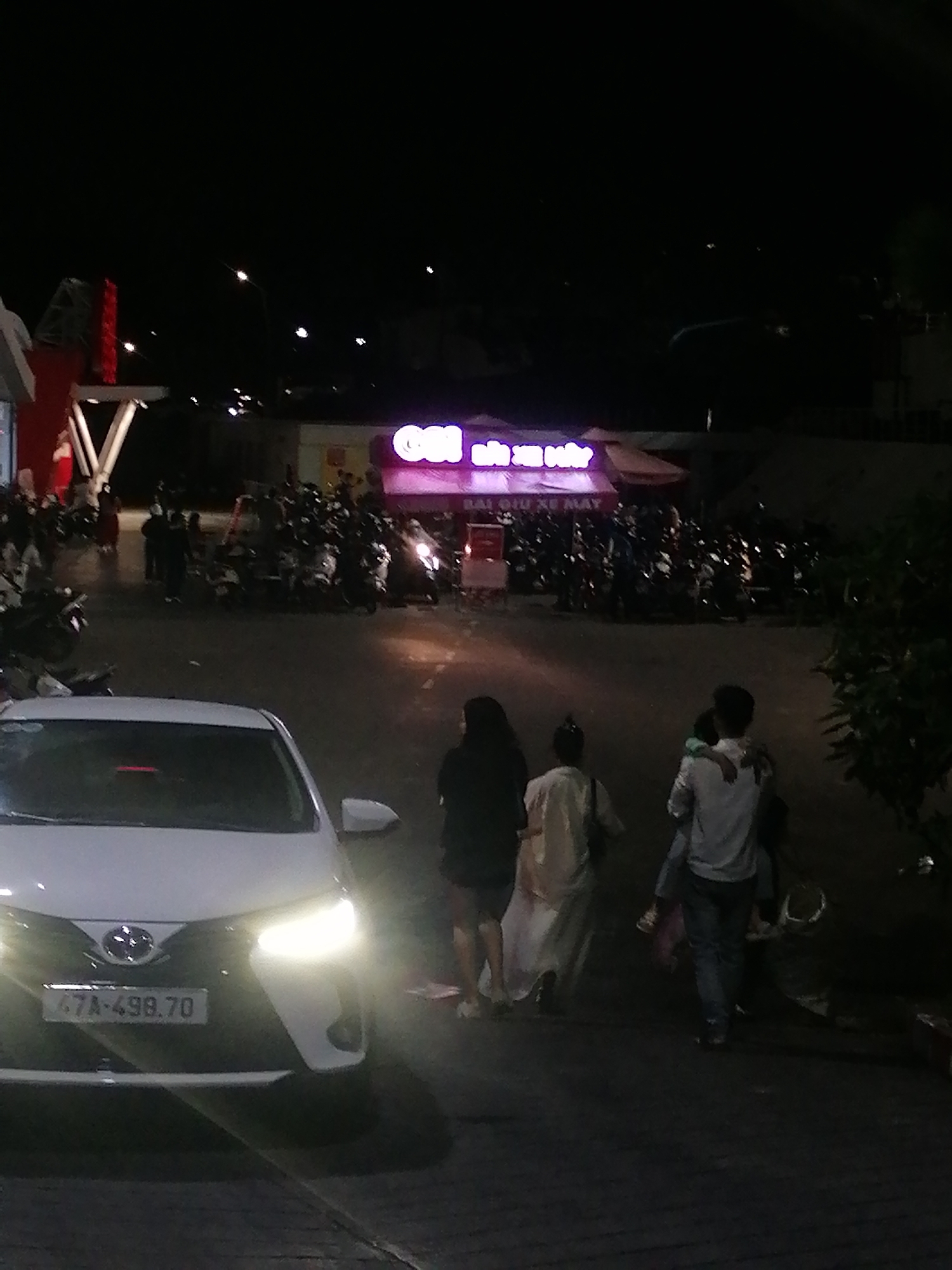 A nighttime scene captures a white car with its headlights on, parked on a paved surface in front of a building with a neon sign that reads "OSA" in large, glowing pink letters. Multiple motorcycles are parked in front of the building, and several people are visible walking around and standing near the motorcycles. A family of four is walking away from the camera towards the building. The father is carrying a child on his shoulders. The mother and another person are walking beside them. The environment appears to be a commercial area in Da Lat, Vietnam, as suggested by the license plate on the car, "67A-498.70," which is a Da Lat license plate. The scene is dimly lit by artificial lights from the building and the car's headlights.