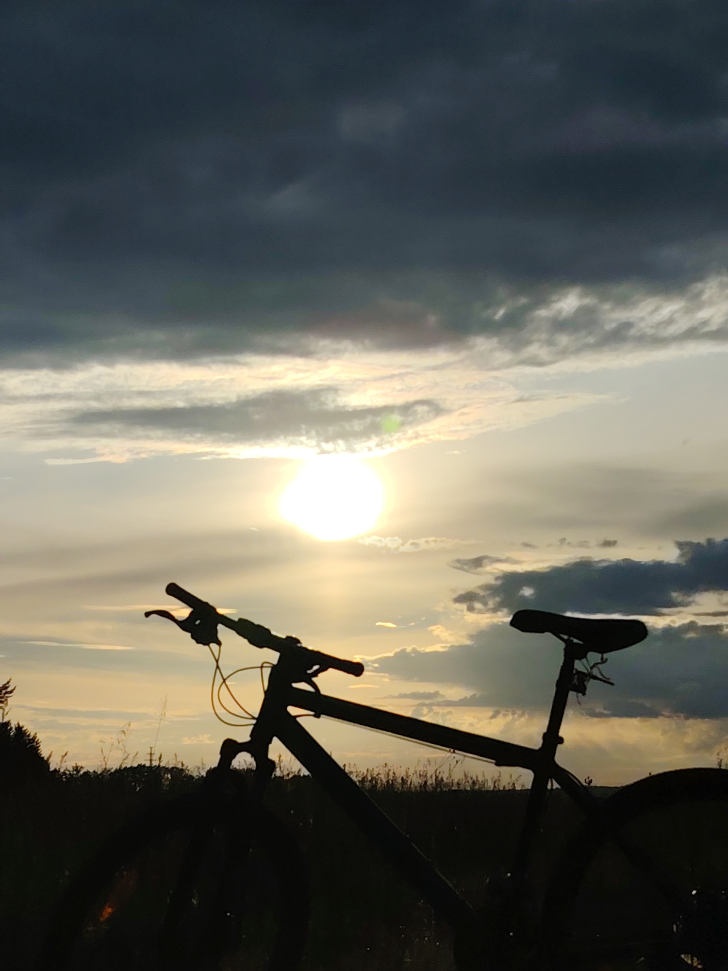 This media file depicts a silhouette of a bicycle against a dramatic sunset sky. The bicycle is positioned in the foreground, with its handlebars, seat, and parts of its frame clearly visible in dark shadow. A yellow wire hangs from the handlebars. The setting appears to be an outdoor, rural location, suggested by the silhouettes of grass and trees along the horizon.
The sky is filled with a mixture of dark, heavy clouds and lighter, wispy clouds illuminated by the setting sun. The sun itself is a bright, glowing orb, partially obscured by clouds, casting a warm, golden light across the scene. The contrast between the dark silhouette of the bicycle and the bright, dynamic sky creates a visually striking image.
The time of day is clearly sunset, with the low angle of the sun and the warm colors in the sky. The weather appears to be mixed, with indications of both overcast conditions (dark clouds) and clearing skies (illuminated clouds).
No people are visible in the image. No text is present. The overall mood of the image is one of tranquility and natural beauty, perhaps evoking a sense of adventure or the end of a day's journey. The location context provided is Chaikovsky, Russia, suggesting this scene is likely from that region.