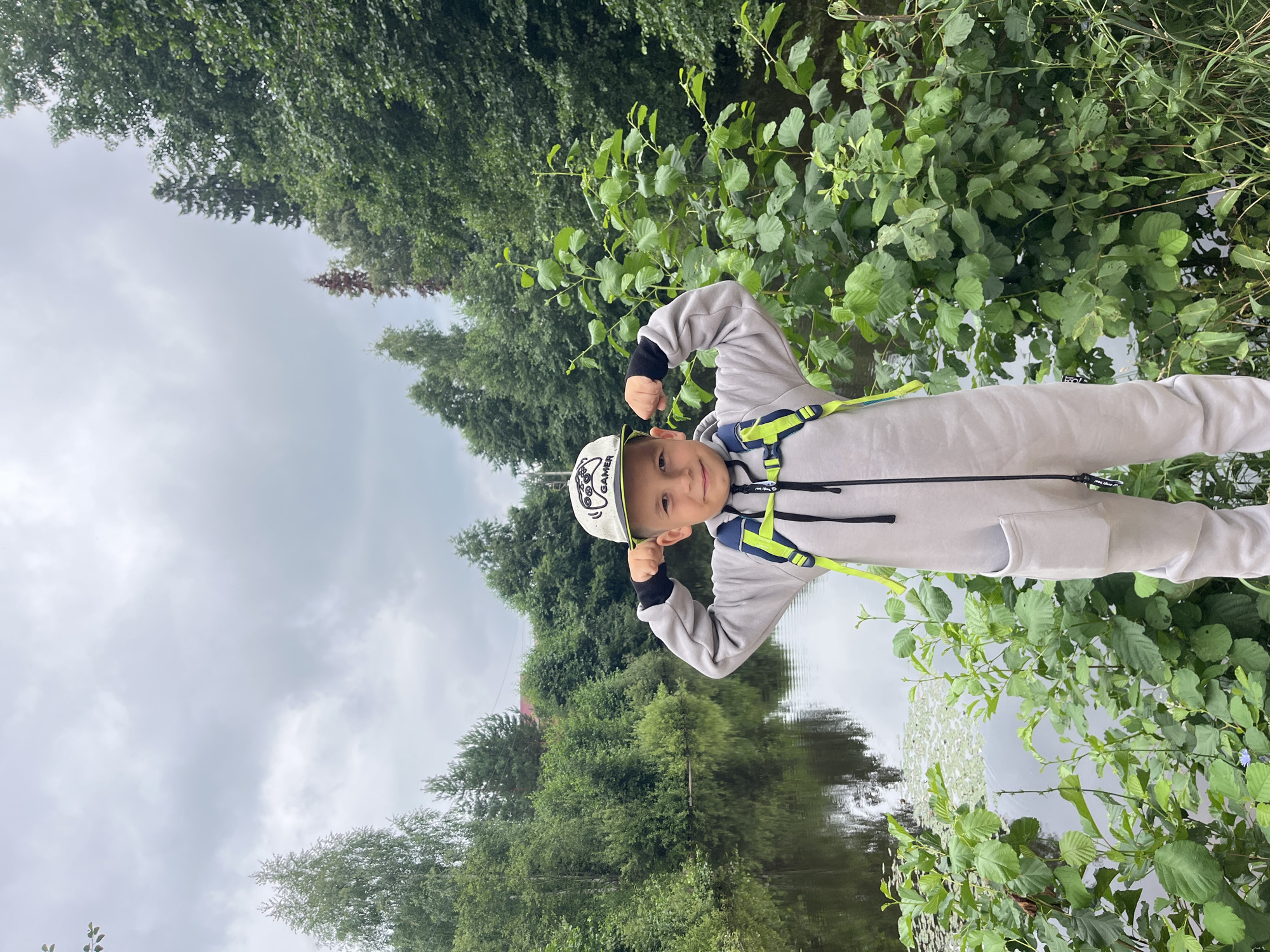A young boy wearing a gray jumpsuit and a cap with "GAMER" written on it, is posing with flexed biceps in front of a body of water. The boy has a backpack with yellow straps. He is surrounded by lush green trees and vegetation. The sky is overcast, suggesting a cloudy day. The location appears to be a natural setting, possibly a park or riverbank in the city of Chaikovsky, Russia, characterized by its abundant greenery and calm water.