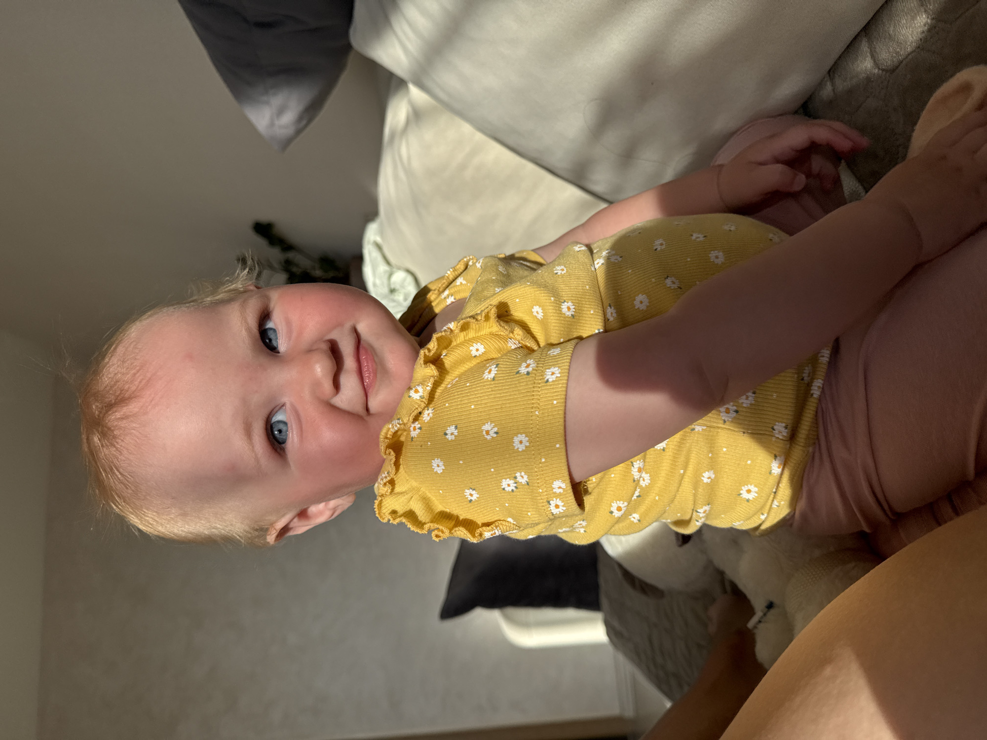 A fair-skinned baby with light red hair and bright blue eyes is depicted sitting upright, looking slightly upward and to the viewer's right, with a subtle smile. The baby wears a ribbed yellow short-sleeve top adorned with white daisy patterns and light pink pants. Strong, directional sunlight illuminates the baby's face, hair, and the right side of the body, casting distinct shadows on the left arm and torso. The setting appears to be a domestic interior, specifically a living room or bedroom, in the city of Chaikovsky, Russia. The baby is seated on a light gray textured surface, possibly a blanket on a couch. To the immediate left, a portion of an adult's bare leg is visible, along with a small white plush object and a slender white stick-like item. The background consists of a plain light-colored wall on the left, partially obscured by a dark gray cushion and a white furniture piece. On the right, a white or cream-colored couch with pillows, including a dark gray one, is visible, with a glimpse of light green fabric further back. The scene is illuminated by natural light, suggesting daytime.