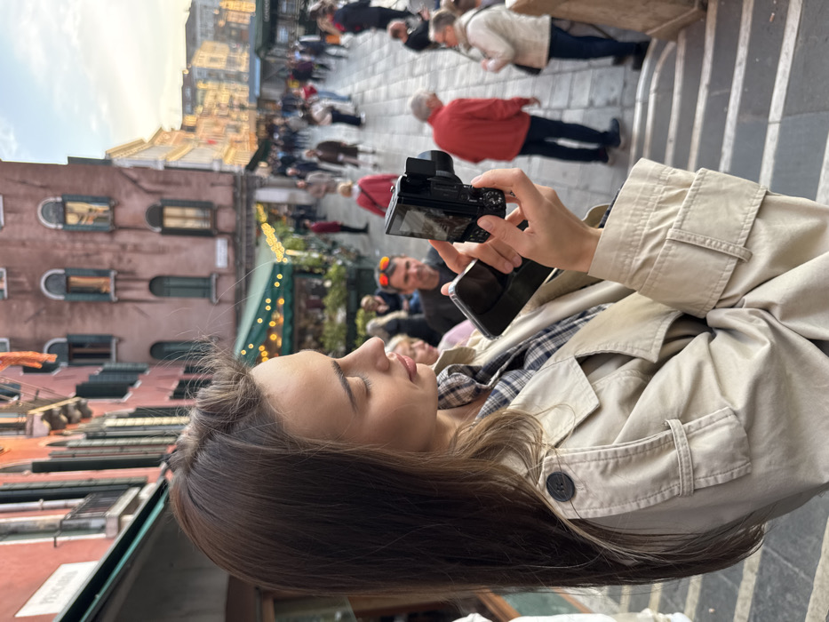 The image depicts a young adult female in the foreground, positioned in profile facing right, within a bustling urban environment in Venice, Italy. She has long, dark brown hair and is wearing a light beige trench coat over a plaid shirt. She holds a black compact digital camera with a flip-out screen in her right hand, while her left hand supports a black smartphone beneath the camera. Her attention is directed towards the camera's screen.
The background features a paved public square or wide thoroughfare populated by numerous pedestrians, estimated between 20 to 30 individuals, engaged in walking and standing. Architectural elements include multi-story historic buildings with facades ranging from reddish-brown to lighter tones, featuring arched windows with dark shutters and visible balconies. Below one building, a commercial establishment with a green awning and string lights is visible. The scene is illuminated by ambient light consistent with late afternoon or early evening. A stone barrier or railing is partially visible in the bottom right corner.
