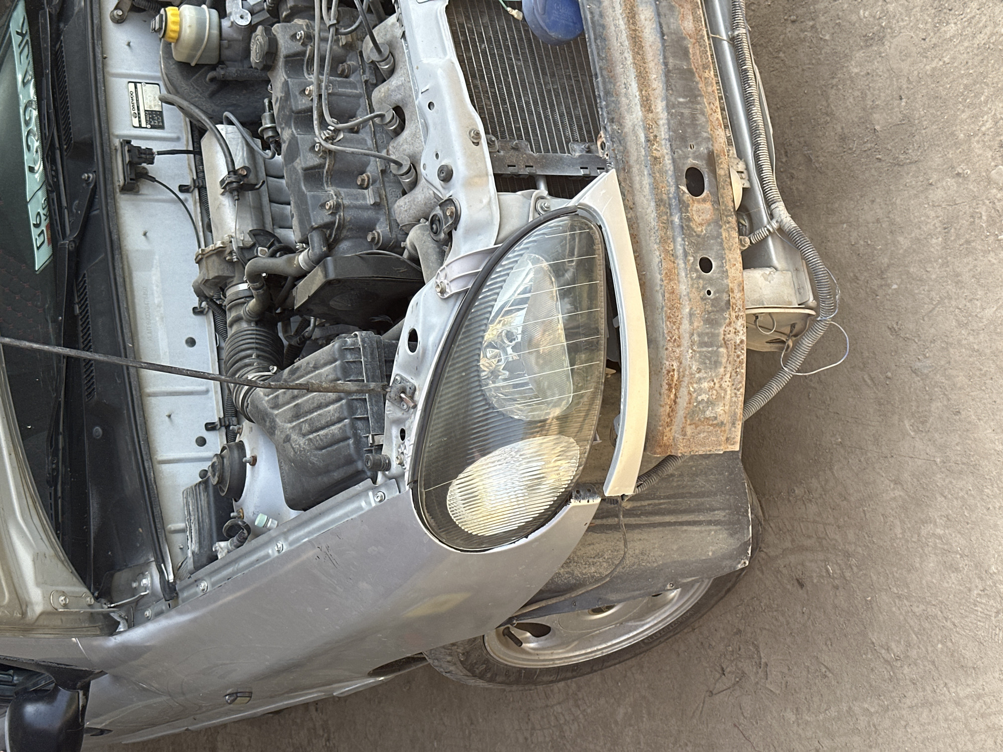 The image shows the front section of a damaged silver car with its hood open, revealing the engine compartment. The car is parked on a dusty, unpaved surface, likely a roadside or a yard. The front bumper is heavily rusted and detached, exposing the car's internal components, including the radiator. The left headlight is intact but the surrounding body panel appears to be slightly misaligned. The tire on the left front is visible and appears to be dirty. The overall condition of the car suggests it has been in an accident or is undergoing repairs. The environment appears to be an outdoor setting in a rural or semi-urban area. There are no people or animals visible in the image. The license plate, partially visible at the top of the windshield, is from Kyrgyzstan.