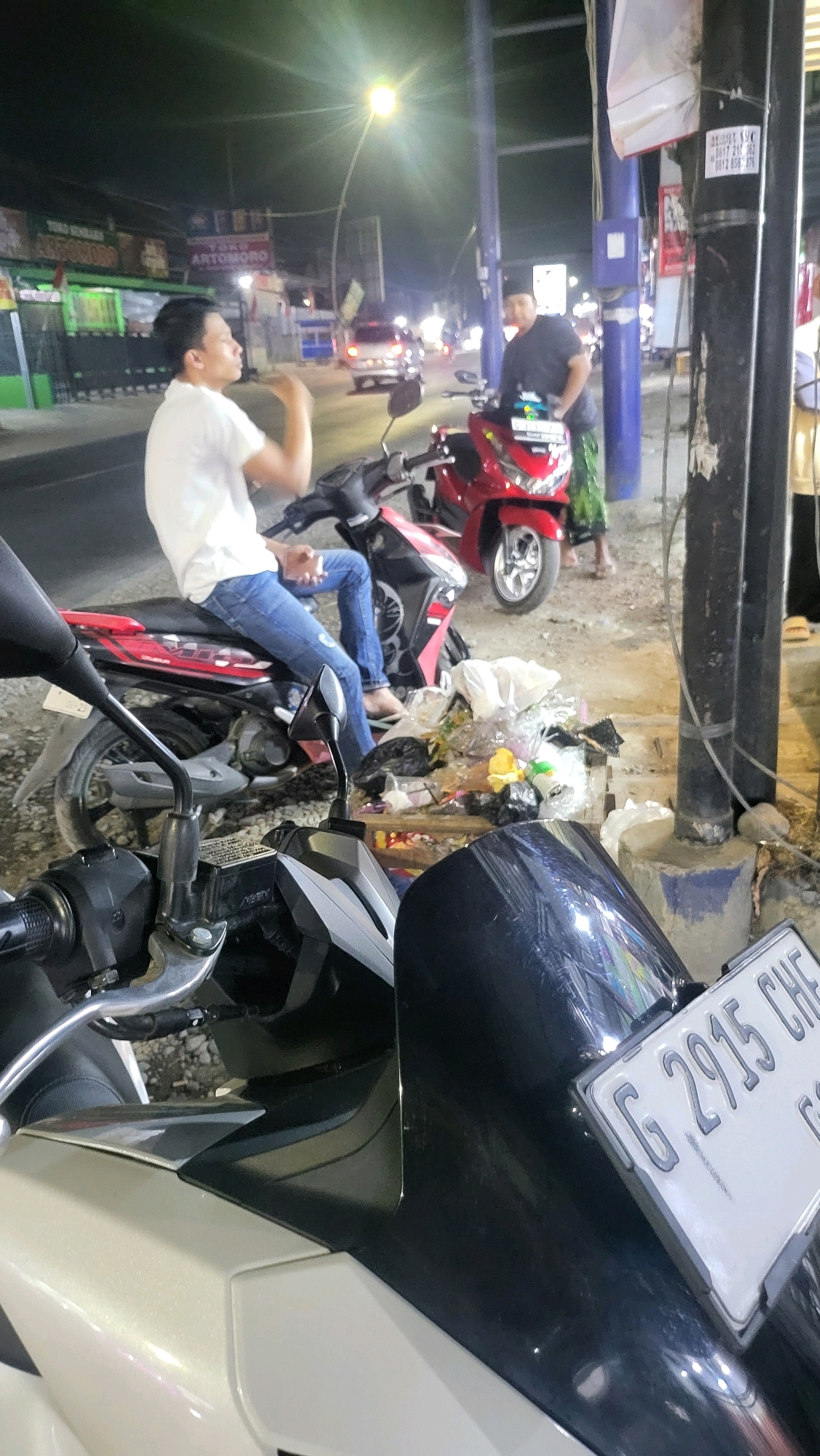 The image depicts a street scene at night in Lebaksiu, Indonesia. In the foreground, a silver and black motorcycle is parked, partially obscuring another motorcycle in the background. Two men are visible. One man, wearing a white t-shirt and blue jeans, is seated on a red and black motorcycle, looking to the left. Another man, wearing a dark shirt and a green and white patterned sarong, stands next to the red and black motorcycle, also looking to the left. A street light illuminates the area, casting shadows. Several bags of trash are piled on the ground next to the motorcycles. In the background, a street lined with businesses is visible, with vehicles and street lights also present. A pole with various signs is situated to the right of the frame. The overall impression is a busy, urban street scene at night, with elements of daily life such as transportation and waste disposal.