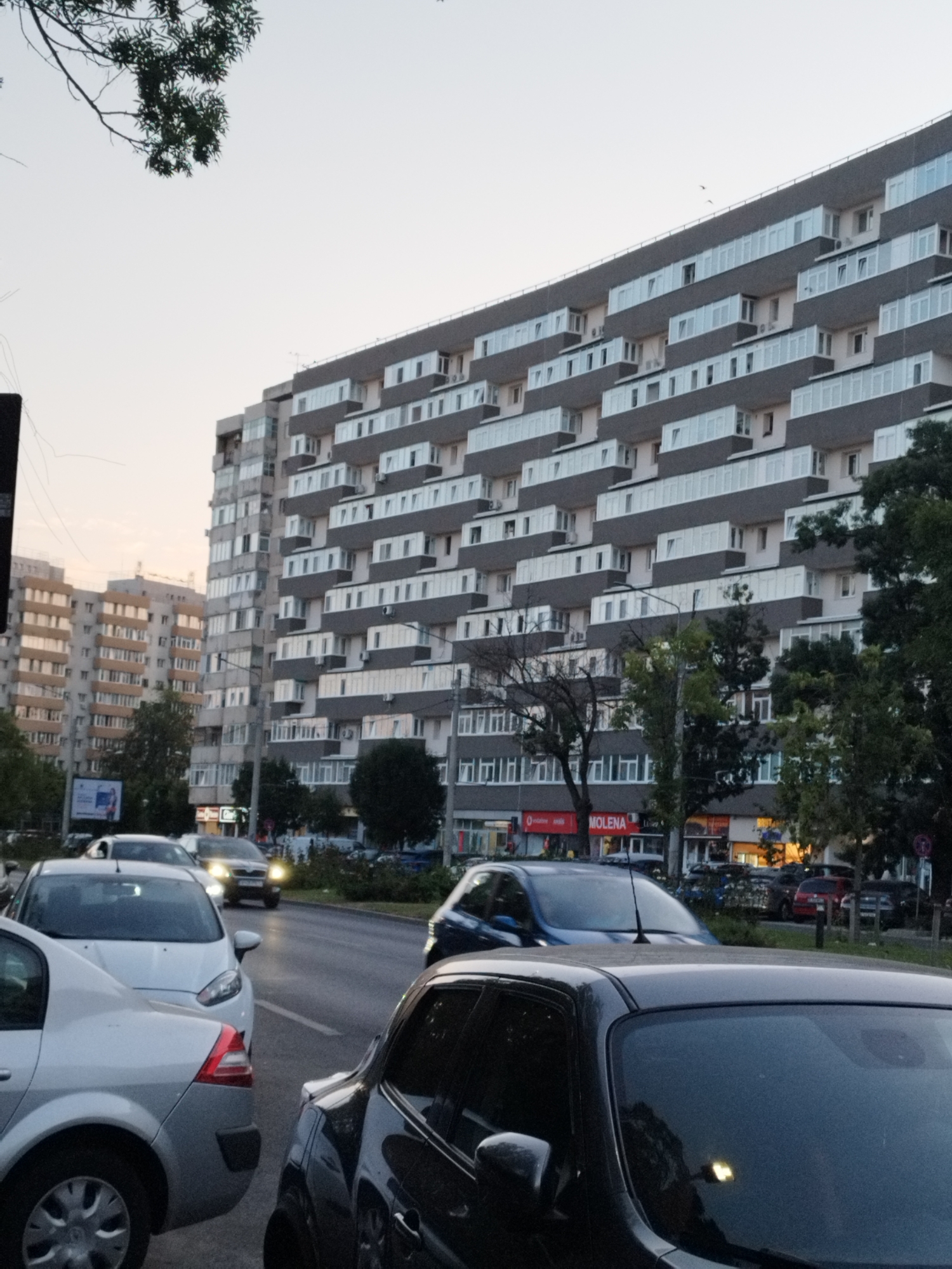 The image captures a street scene in Bucharest, Romania, characterized by dense urban architecture and traffic. Dominating the background is a large, multi-story apartment building with a distinctive facade featuring numerous staggered balconies, each enclosed by glass. To the left, another apartment building, also multi-story, is visible, with a warmer-toned facade. The foreground and middle ground are filled with vehicles, indicating a moderate level of traffic congestion. Cars of various makes and models are present, some in motion and others likely stopped or moving slowly. Trees and foliage are interspersed throughout the scene, suggesting a mix of urban development and green spaces. Storefronts with signage are visible along the street level of the buildings, hinting at commercial activity. The lighting suggests it is either early morning or late evening, with a soft, diffused light. There are no discernible people in the image. The overall impression is of a typical, busy city street in Bucharest.