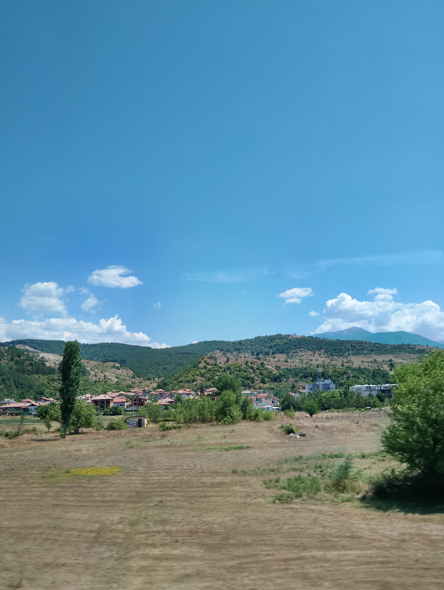 The image depicts a rural landscape with a village nestled in a valley. The foreground consists of dry, golden-brown grass. In the mid-ground, a cluster of red-roofed houses forms a village, with a tall, slender green tree standing prominently to the left of the village. Beyond the village, rolling hills covered in green trees and sparse vegetation rise up, transitioning into more densely forested slopes. The sky is a clear, bright blue with scattered white clouds. In the distance, the outlines of mountains are visible. The scene is captured from a slightly elevated perspective, suggesting the viewer is on a slight incline or looking out from a higher vantage point. There are no people or animals visible in the frame. The overall impression is one of a serene, sunny day in a picturesque countryside.