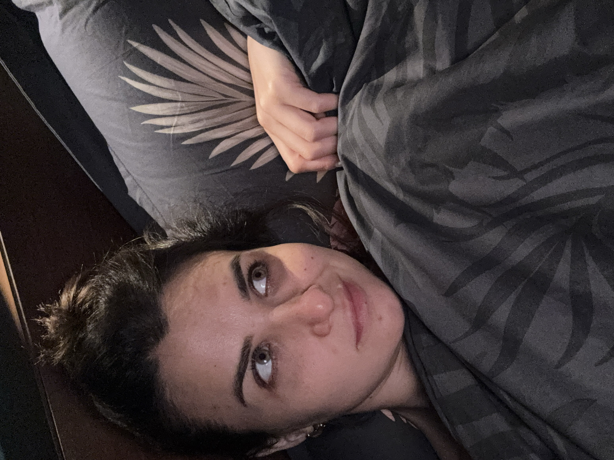 A woman lies in bed, covered partially by a patterned gray blanket. Her head rests on a matching patterned gray pillow with a leaf design. She looks upward with a slight smile. The scene is indoors, likely in a bedroom. There is no information in the image to suggest this is in Tivat, Montenegro.