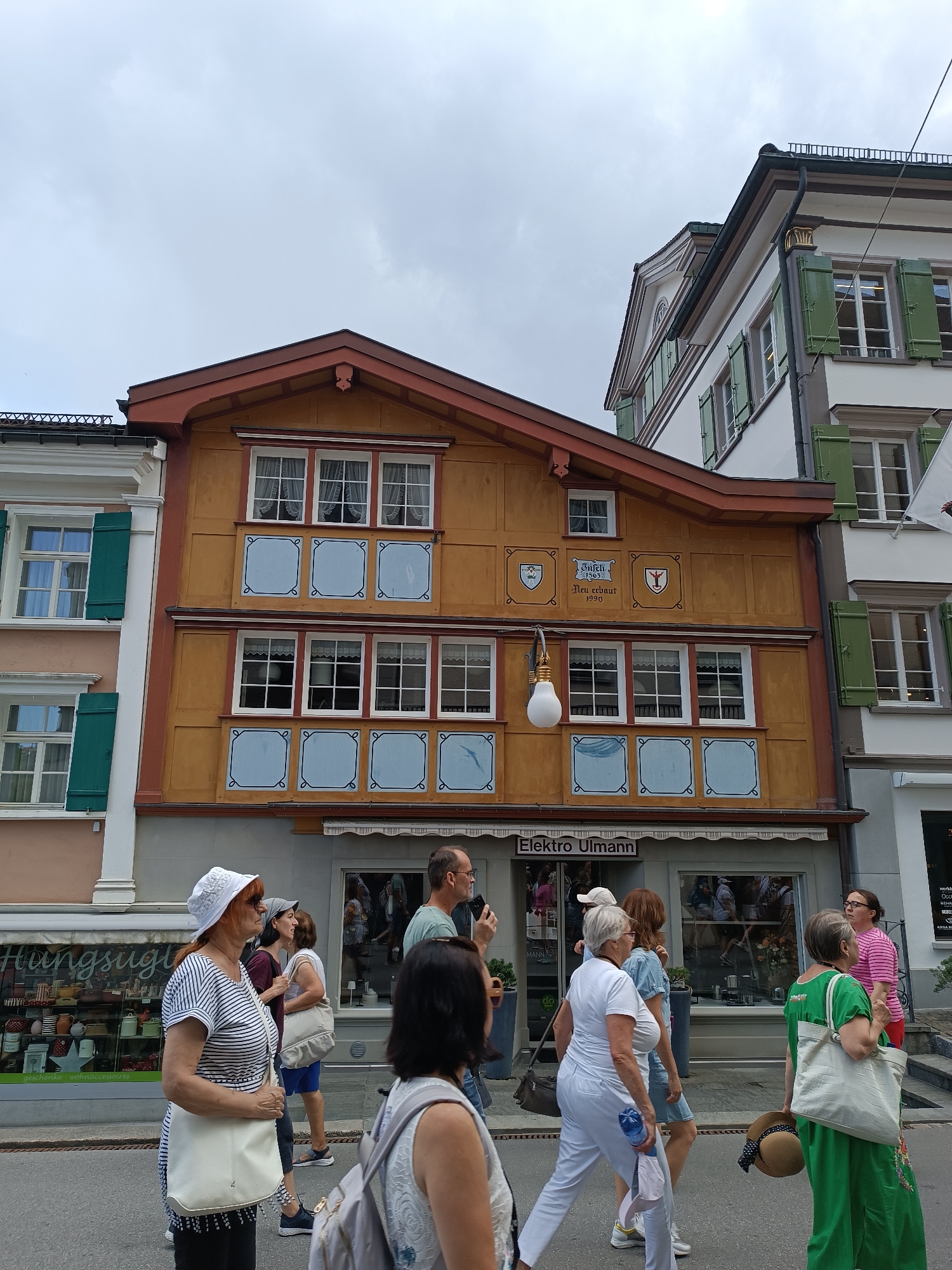 The image depicts a street scene in the city of Appenzell, Switzerland. The central focus is a traditional wooden building with a distinctive orange facade, adorned with decorative blue squares above the windows. Above these squares, there are crests and inscriptions, one reading "Töchti 1853 Neu erbaut 1990". A quaint hanging lamp, resembling a large lightbulb, is suspended in front of the building. Below the windows, a shopfront with the name "Elektro Ullmann" is visible, along with a striped awning.
Several people are walking on the street in front of the buildings. On the left, a woman wearing a striped shirt and a white hat is walking towards the right. To her right, another woman in a dark top and shorts is walking. Further to the right, a group of people are walking in the same direction. One woman is wearing a white top and white pants, another in a blue dress, and a third in a green outfit holding a straw hat. The background shows other buildings with green shutters and cloudy skies. The street appears to be paved.