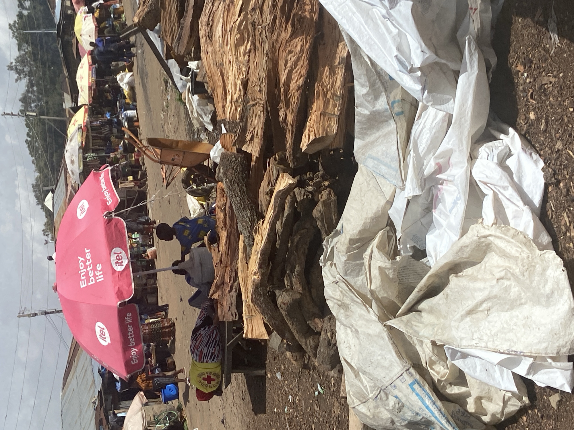 This media file depicts an outdoor market scene in Bukuru, Nigeria. Several people are present, engaged in various activities. In the foreground, a large pink umbrella with the "itel" logo and the text "Enjoy better life" is prominently displayed. A person wearing a yellow vest with a red cross emblem is visible.
The scene appears to be a bustling marketplace, with stalls and goods set up along a dirt path. In the background, other market stalls with umbrellas are visible, suggesting a variety of vendors and products. Piles of raw timber or firewood are stacked in the middle ground, a significant visual element in the image. Several white plastic bags or tarps are also present, some appearing to be stored or discarded. A wheelbarrow is visible near the timber. The overall impression is one of everyday commerce and activity. The time of day appears to be daytime, with sunlight casting shadows, and the weather seems fair. The ground is unpaved, typical of many market areas.