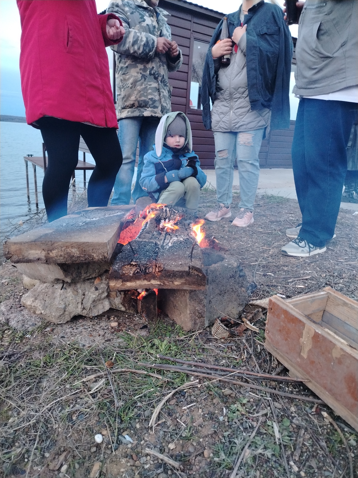 The image captures an outdoor scene in Chaikovsky, Russia, featuring a group of people gathered around a campfire by a large body of water. In the foreground, a small child, dressed in a blue jacket, green pants, blue gloves, and a grey hooded hat with distinctive bunny ears, sits directly in front of a roaring campfire. The fire is built on the ground, contained by large wooden logs or concrete blocks.
Several adults are partially visible around the fire. On the left, a person wearing a bright red jacket and black leggings stands near the water. Beside them, another individual is seen in a camouflage jacket and blue jeans. To the right of the child, a person in a grey quilted jacket, light blue ripped jeans, and a dark denim jacket draped over their shoulders holds a dark bottle. Another person in a grey jacket and blue jeans stands further right. All individuals are dressed in warm attire, suggesting cool weather or an evening setting.
In the background, a wooden building, possibly a cabin or shed, is visible. A simple wooden bench or table stands near the water's edge, partially submerged. The sky above is overcast. The ground is a mix of dirt, grass, and gravel, with scattered twigs and a wooden crate-like object on the right side of the frame. The scene portrays a casual gathering, likely for warmth and companionship around the campfire. There is no visible text in the image.