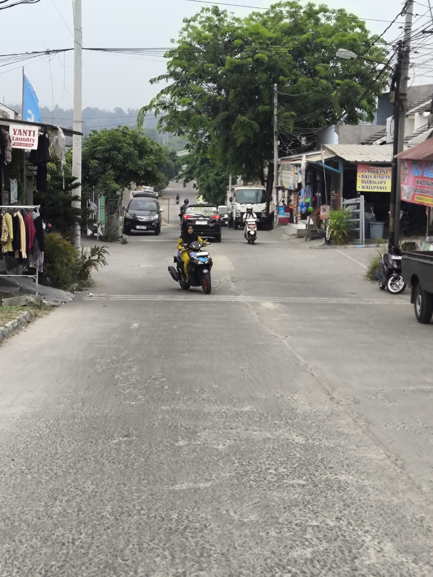 The image depicts a street scene in Cilegon, Indonesia, likely in a commercial or residential area. The foreground shows a grey asphalt road with some texture and patches of wear. In the middle ground, several vehicles are present. A woman wearing a yellow hijab and outfit is riding a black and blue scooter, moving towards the viewer's right. Behind her, a black SUV is stopped on the road. Further down the street, another black SUV is also stopped, and a white truck is visible. On the left side of the street, a laundry business named "YANTI LAUNDRY" with a blue flag is visible, with clothes hanging on a rack outside. Shops with awnings are present on the right side of the street, with signs indicating businesses. Trees with green foliage are scattered throughout the scene, and utility poles with numerous wires are prominent in the sky. The overall atmosphere is overcast, suggesting a cloudy day.