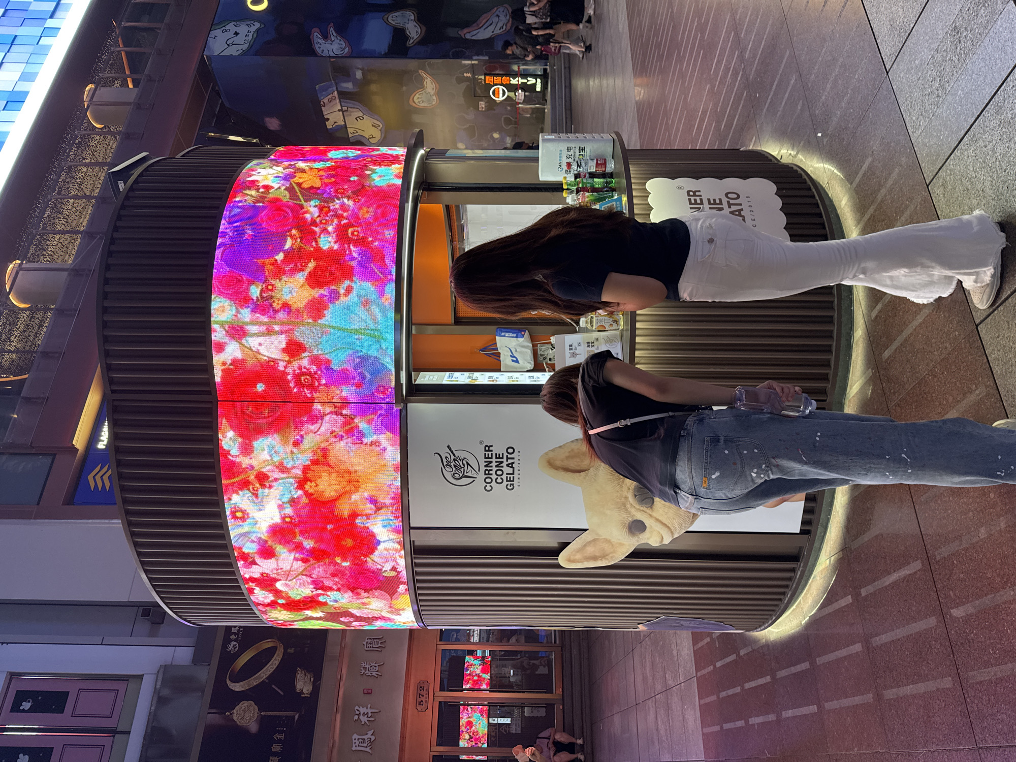 Two young women stand at a brightly lit gelato stand in a bustling city environment, likely Shanghai, China. The stand, branded "CORNER CONE GELATO," features a large, vibrant LED screen displaying a floral pattern. One woman, in the foreground and facing away, wears a dark t-shirt and distressed denim jeans with paint splatters, holding a clear plastic bottle. The second woman, to her right and also facing the stand, is dressed in a dark t-shirt and white flared pants. They appear to be interacting with the menu or ordering. The background shows more urban elements, including storefronts with illuminated displays and other pedestrians. The ground is paved with reddish-brown tiles, and the overall lighting suggests it is evening.