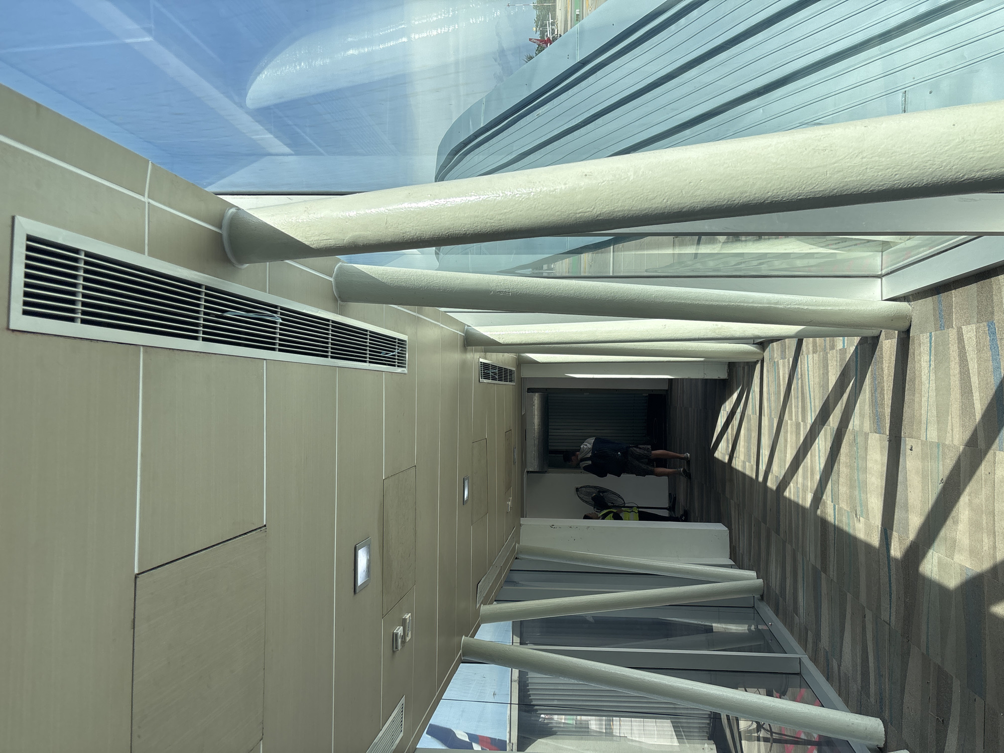 This image depicts the interior of a jet bridge or airport walkway at Phuket International Airport (HKT) in Mai Khao, Thailand. The perspective is from within the walkway, looking towards its far end.
The ceiling consists of light-colored rectangular panels, some showing minor discoloration or misalignment. Integrated into the ceiling are several rectangular recessed light fixtures and long, gridded rectangular air conditioning vents.
The floor is carpeted with a patterned design composed of horizontal stripes in varying shades of grey, blue, and beige. Direct sunlight streams through the windows on the right, casting prominent, elongated diagonal shadows across the floor.
Along the right side of the walkway, a series of thick, cylindrical support columns, painted a light, textured off-white color, are angled inward from the base to the ceiling. These columns stand between a full-height glass window wall that runs the length of the walkway.
Through the window, an external airport environment is visible. A large, curved architectural structure with a ribbed or corrugated metallic surface, possibly a roof or part of another terminal building, occupies a significant portion of the outdoor view. Beyond this structure, a blue sky with some cloud cover is apparent. Further in the distance, parts of a red and white aircraft, along with a light pole, are discernible on the tarmac.
Towards the far end of the walkway, two individuals are present. One individual, closer to the center, is partially obscured but appears to be wearing a high-visibility vest and is near a black portable fan positioned on the floor. The second individual, further down the walkway, is seen from behind, wearing a dark hooded top, shorts, and a backpack, and appears to be standing or walking away from the viewer. The far end of the walkway leads to an interior area with a solid wall.