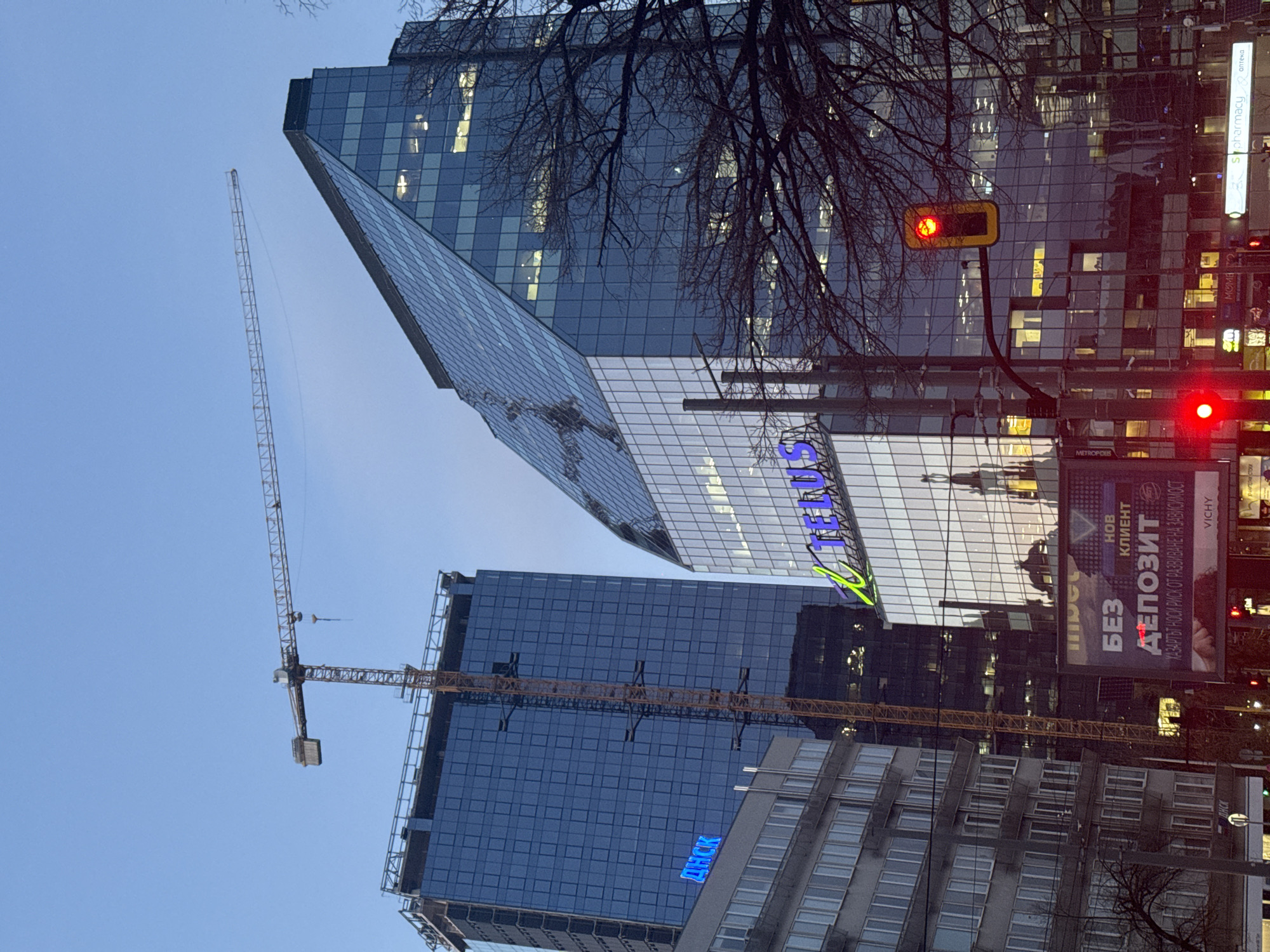 The image captures a street view in Sofia, Bulgaria, at dusk or early evening. The sky is a pale blue. Two tall, modern glass-clad buildings dominate the skyline. The building in the foreground is still under construction, with cranes visible against the sky. The second glass clad structure has the text "Telenor" in blue. In the immediate foreground is a red stop light. A sign with Russian text (ДНСК, БЕЗ, ДЕПОЗИТ) and some advertising is to the right side of the scene, along with light poles. The street is lined with other, shorter buildings, and some bare tree branches obscure parts of the scene.