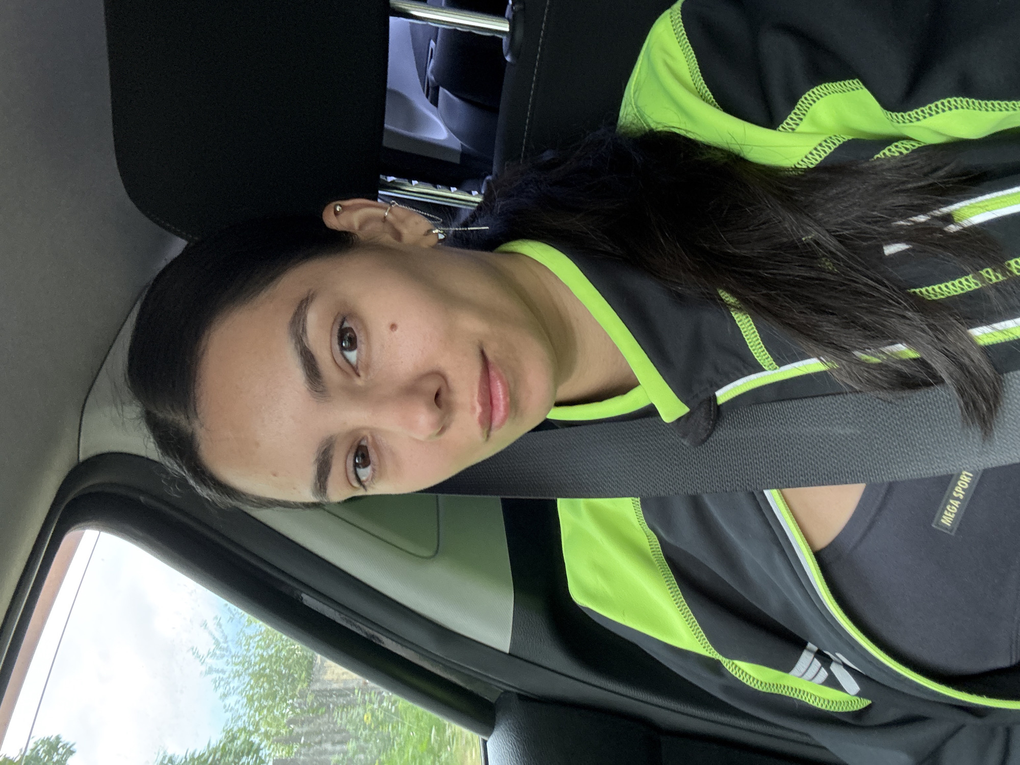 A woman sits in the driver's seat of a car, wearing a seatbelt, a black top with a neon green stripe, and various earrings in her left ear. The car window shows a view of trees. The car's dashboard and interior are visible. Based on the presence of the words "MEGA SPORT" on her top, it is possible that the photo was taken in or related to an area of sporting activity or commerce. Given the prompt's inclusion of Chaikovsky, Russia, it is presumed the location is within that city, although the image itself does not directly confirm that.
