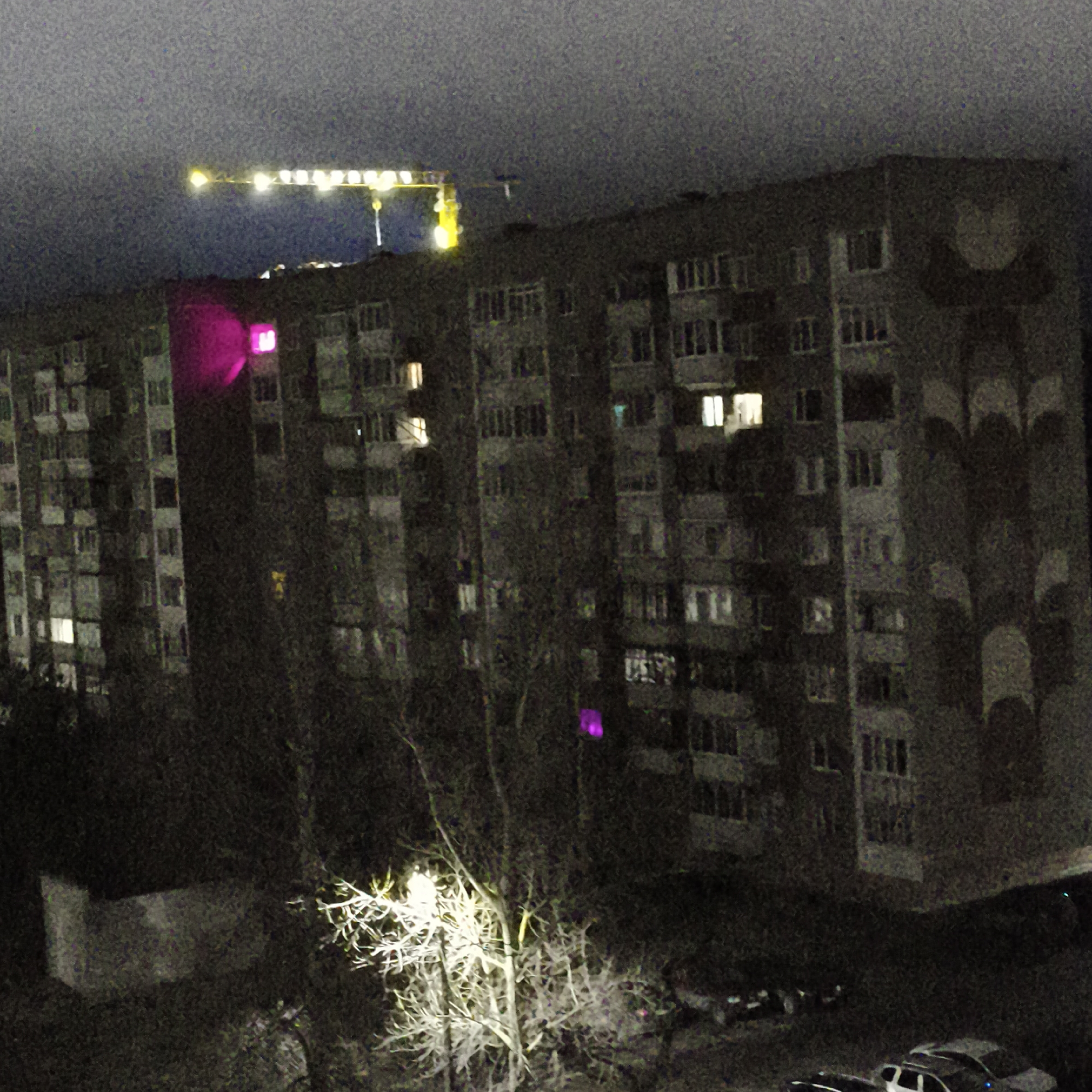 At night in Chaikovsky, Russia, a low-angle shot reveals a multi-story apartment building, with a construction crane visible behind it, its lights illuminating the sky. The building is illuminated by various lights, including at least one purple light. In the foreground, a barren tree stands illuminated, with parked cars underneath. A wall appears in the left foreground. The scene is dark and grainy.