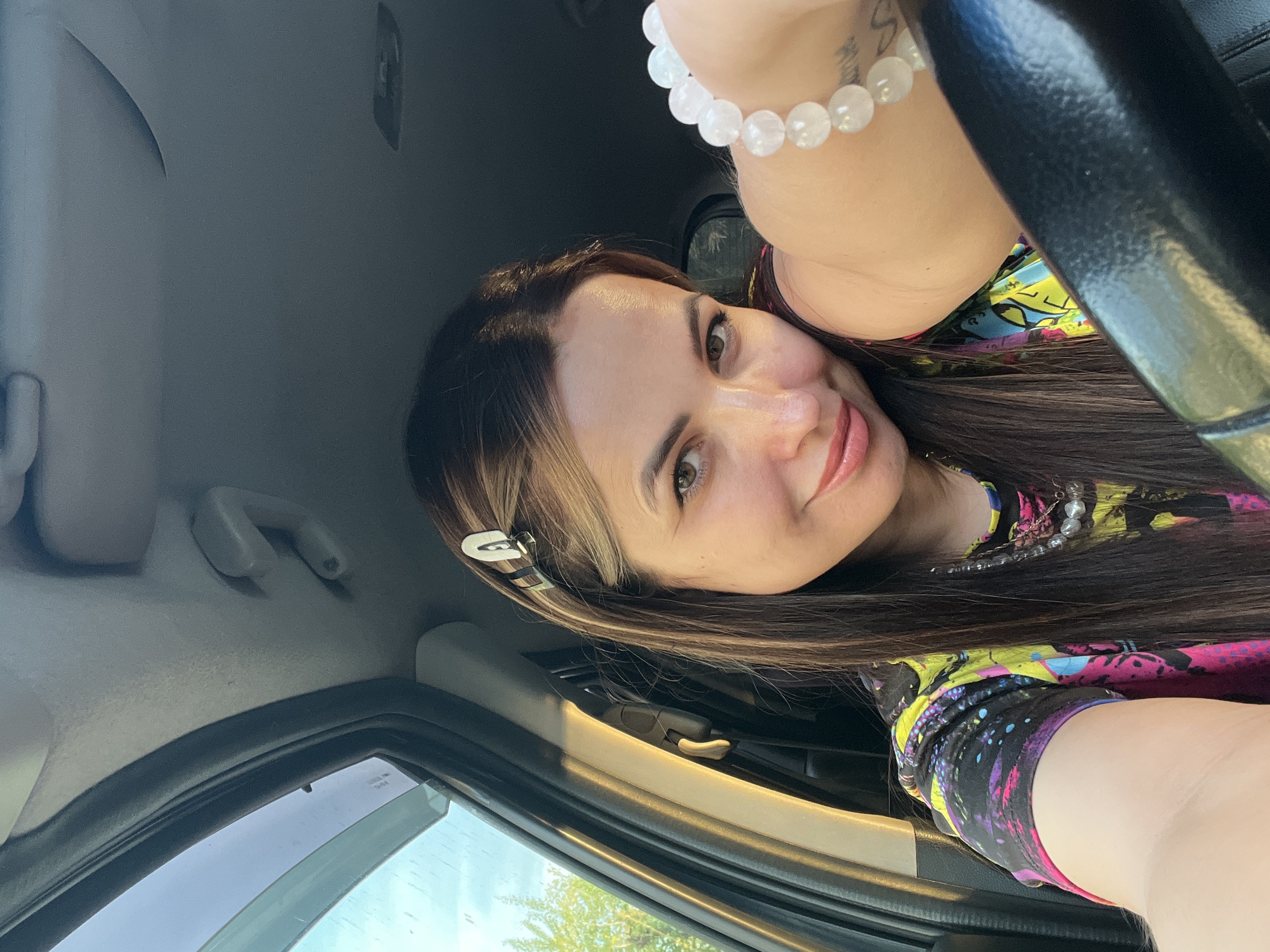A woman with long, brown hair and a patterned top is in the driver's seat of a car in Unknown, Unknown. She is smiling and has a white bracelet on her wrist. Her hair is styled with a silver clip. The car's interior is visible, including the steering wheel, window, and grab handle. The background shows a glimpse of trees outside the car window.
