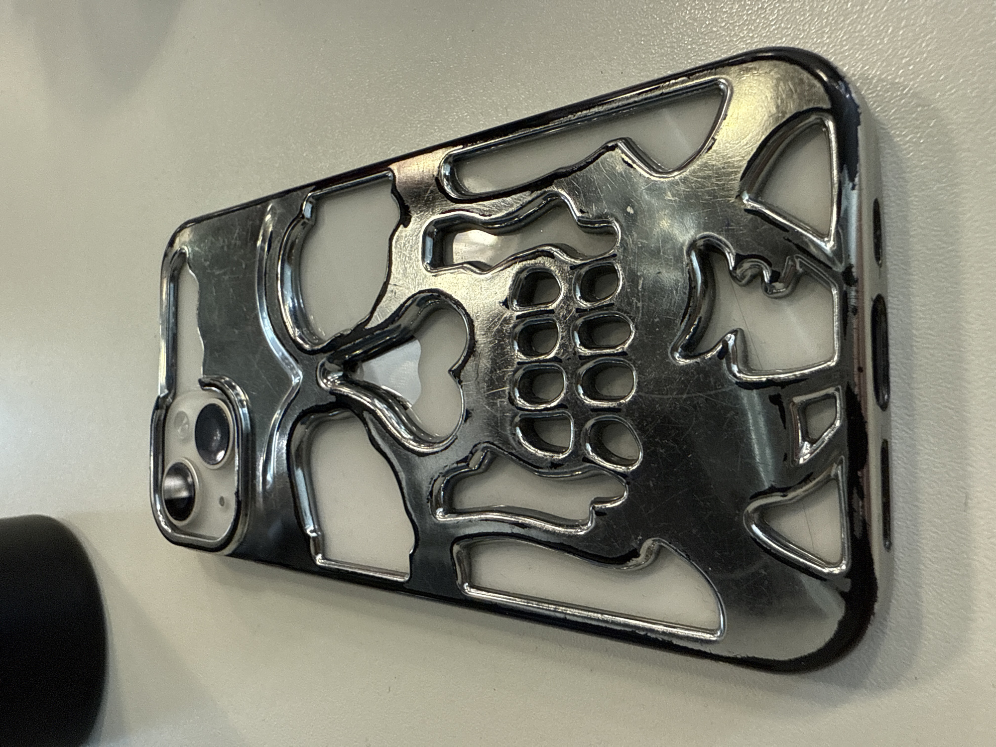 The image shows a close-up, high-angle shot of a chrome-finished iPhone case with a skull design. The case is lying on a light-colored, textured surface, likely a desk or table. The skull design is intricate, with cutouts for the eyes, nose, and teeth, giving it a skeletal appearance. The chrome finish is reflective, showing some scuff marks and fingerprints, suggesting it has been used. In the upper left corner of the image, a blurred dark object, possibly a water bottle or a mug, is partially visible. The background is plain and out of focus. There are no people present in the image, and no specific urban or natural environment details are visible, making it impossible to determine a location in Perm, Russia, or any other city.