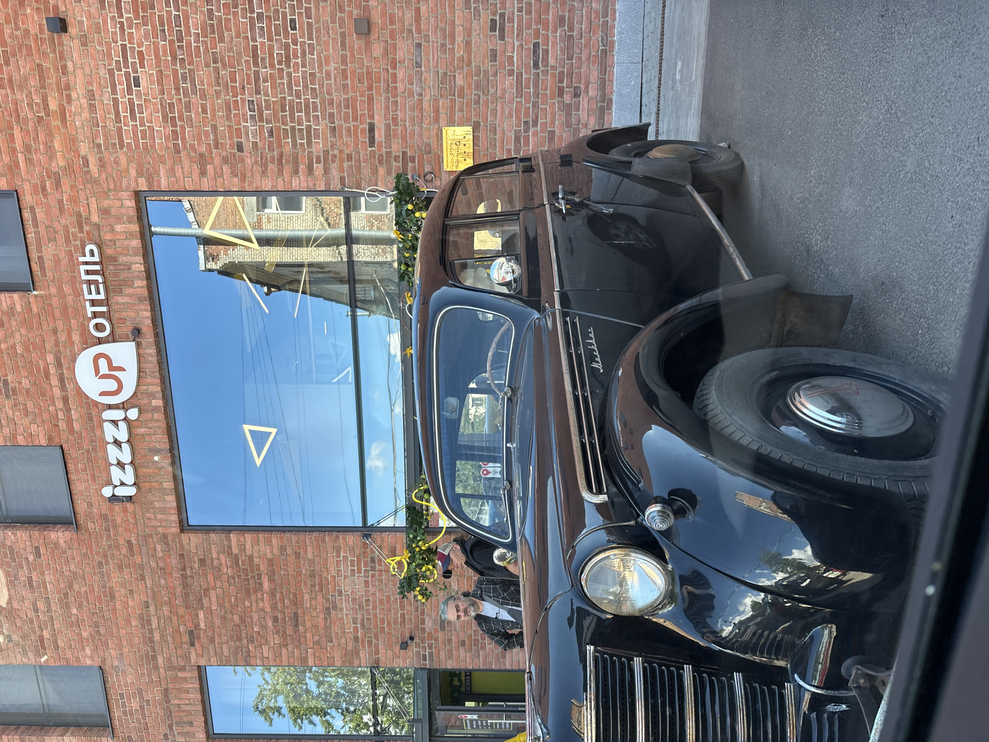 An old black Moskvich car is parked in front of a brick building with large windows. The building has a sign that reads "izzzi UP ОТЕЛЬ". The car has chrome trim and a distinctive front grille. Two men are standing near the car, with one leaning against the building and the other sitting in the driver's seat. The scene appears to be in Saint Petersburg, Russia, as indicated by the architecture and the Cyrillic lettering on the sign. The overall impression is a juxtaposition of vintage automotive charm and modern urban architecture.